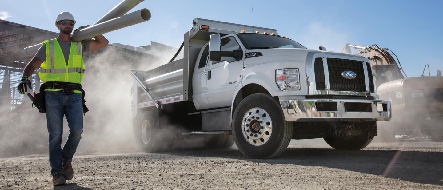 Worker carrying pipes on a dusty construction site next to a 2026 Ford F-750® with dump truck upfit shown in Oxford White
