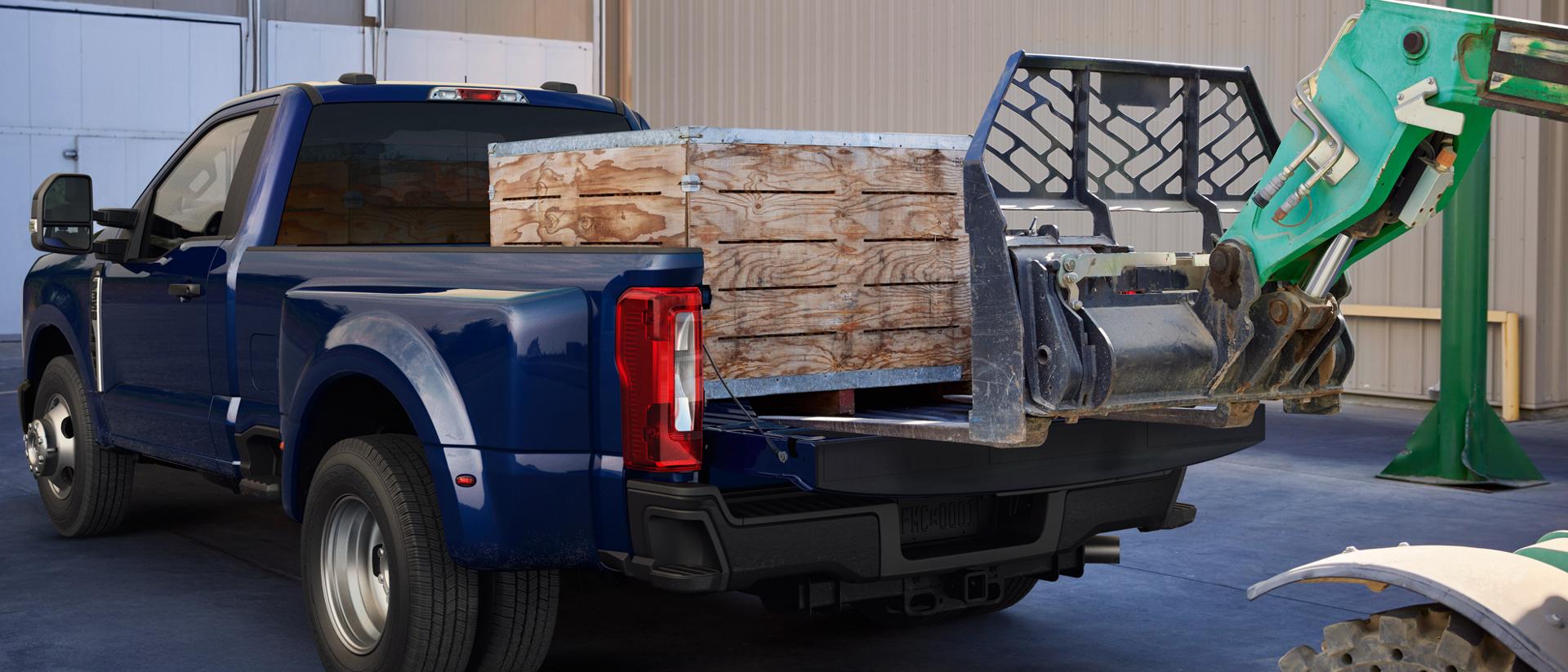 A 2026 Ford Super Duty® F-350® Lariat® model in Argon Blue with a heavy crate being loaded into the truck bed