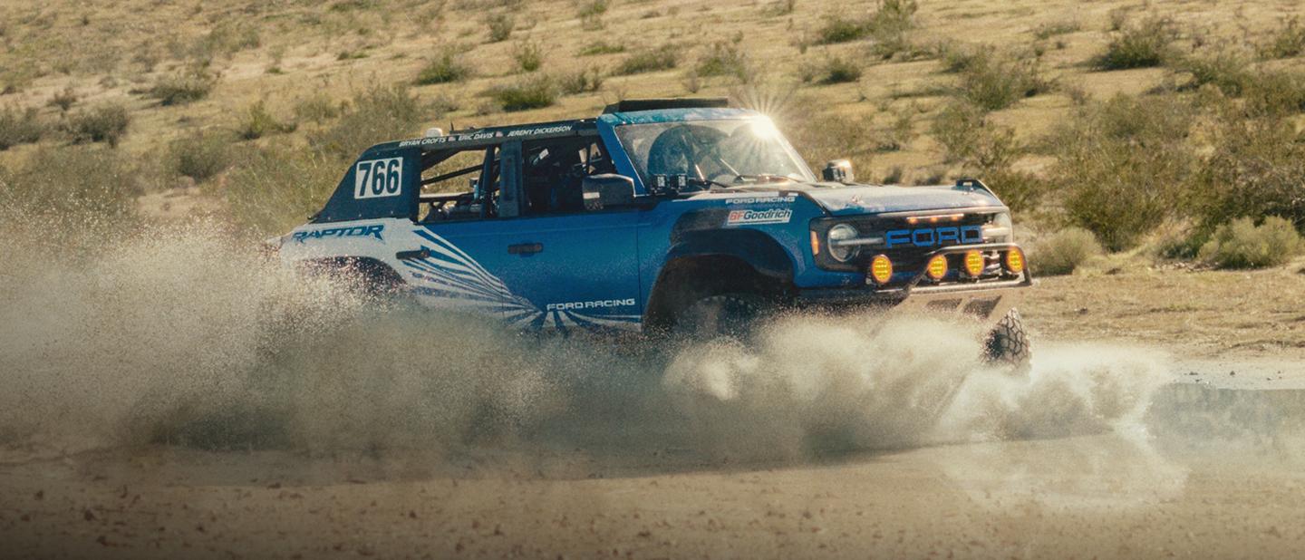 A blue with white graphics 2025 Ford Bronco® Raptor® racing through the desert