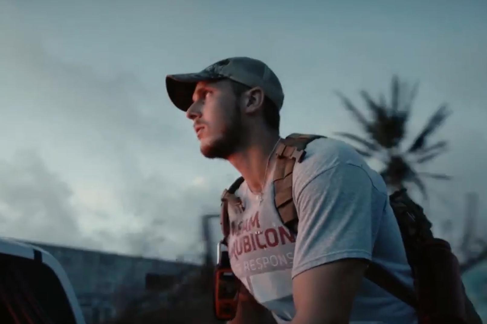A person wearing a backpack and baseball cap sits in the bed of a truck