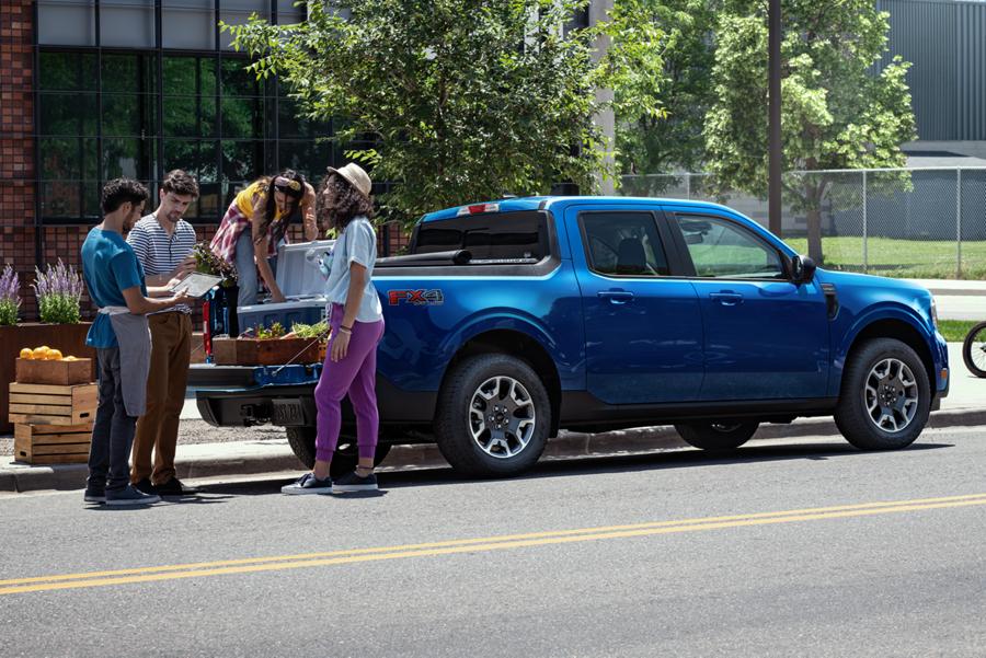 A group of people loading plants into the bed of a blue Ford Maverick pickup truck that's parked at the side of a city street