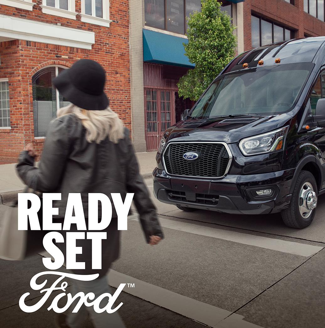 A three-quarter front view of a 2025 Ford Transit driving in an urban setting. In the lower left hand corner white text appears, “Ready Set Ford”. 