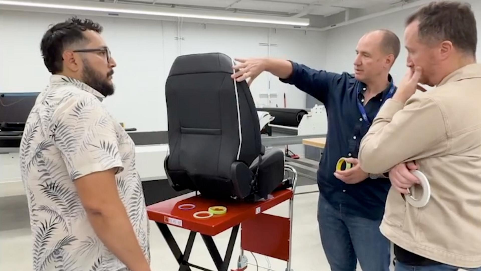 Three men standing around a seat discussing the materials