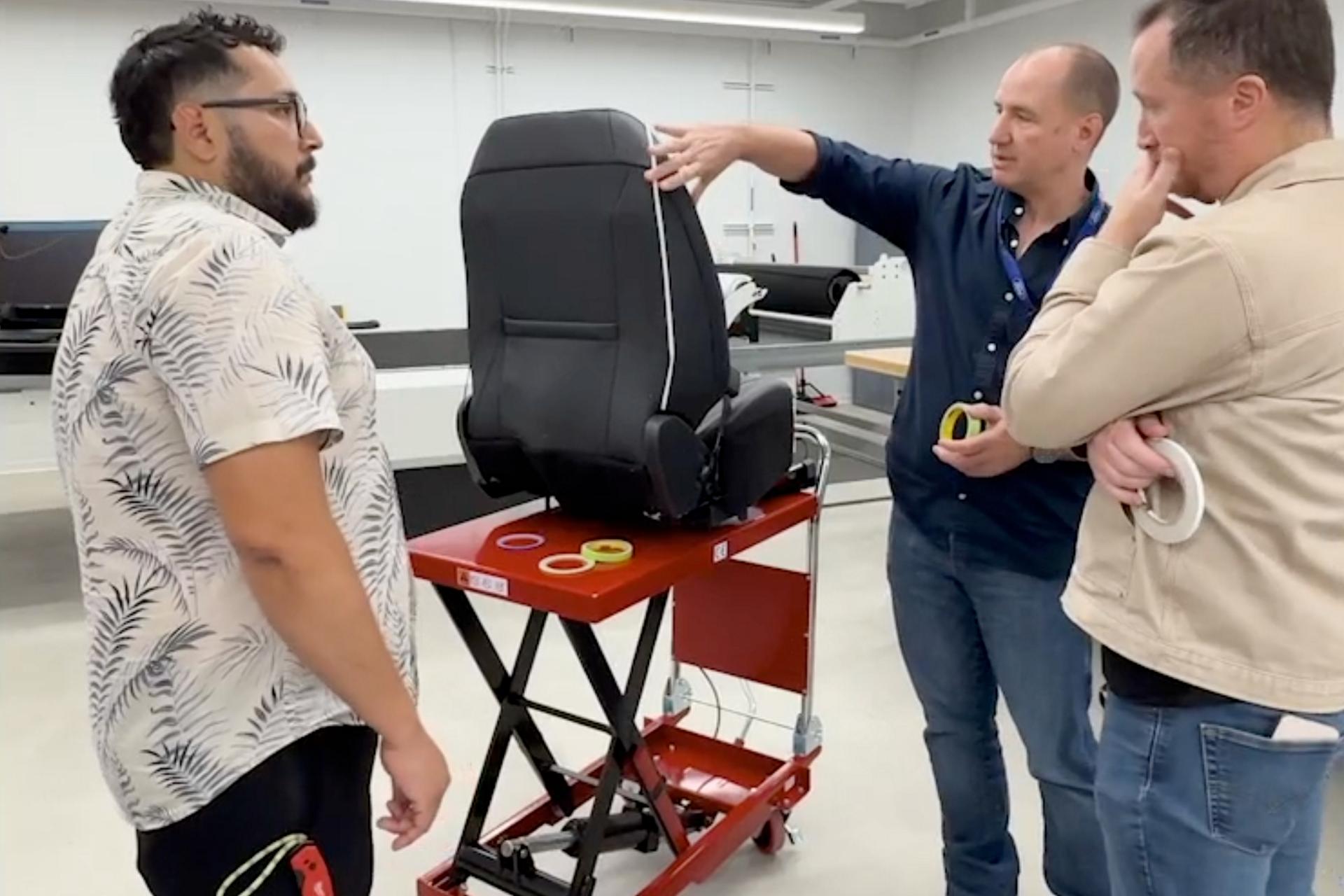 Three men standing around a seat discussing the materials