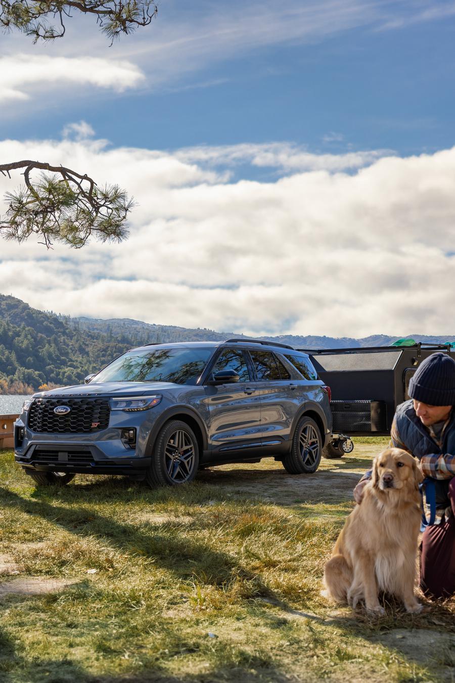 A 2025 Ford Explorer® ST SUV parked near the edge of a lake with a dog and family setting up a tent at their camp site
