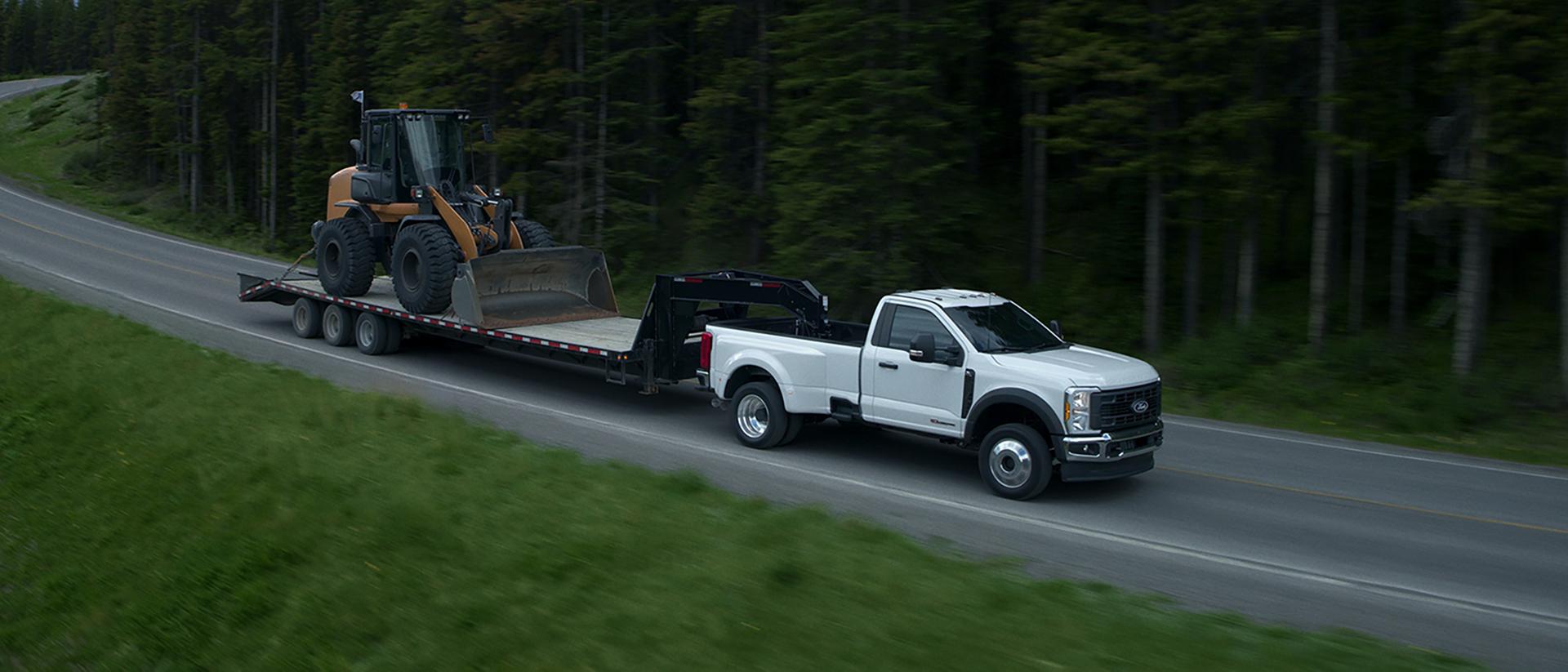 A white F-Series truck towing an industrial digger on a trailer down a forest-lined highway