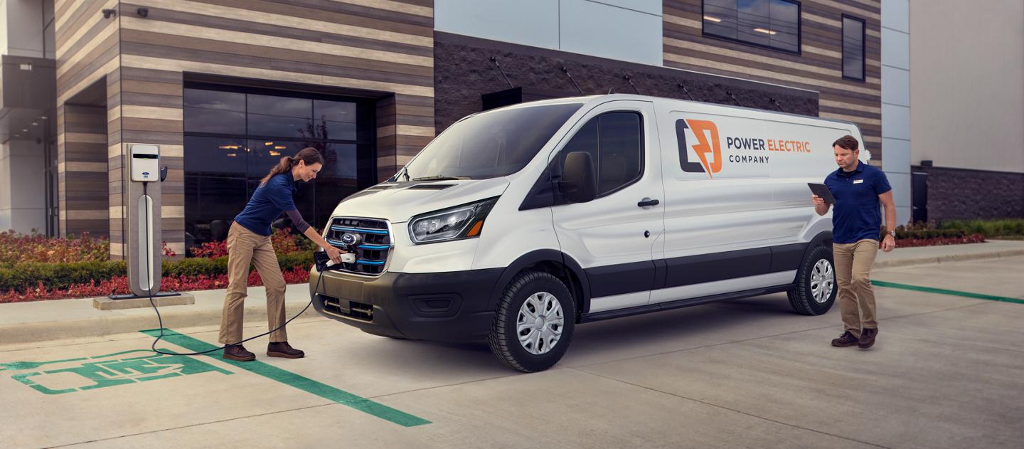 A white 2025 Ford E-Transit™ van parked and being charged by a worker outside a business