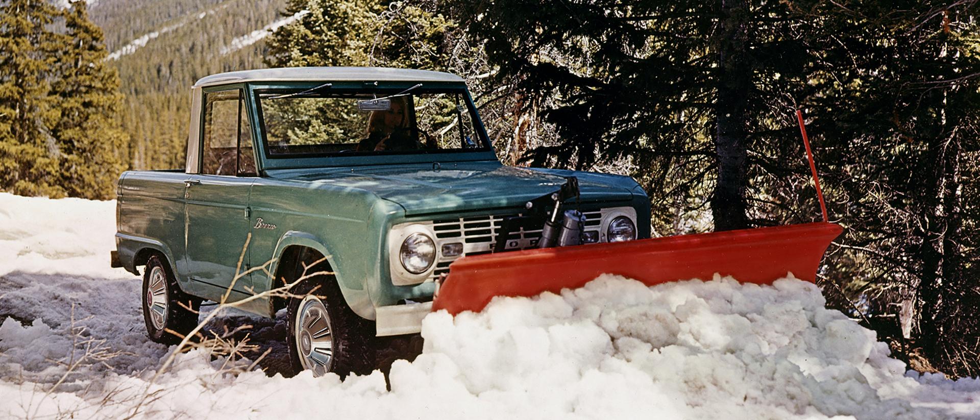 A 1967 Ford Bronco® model equipped with a front snowplow clearing a forest road