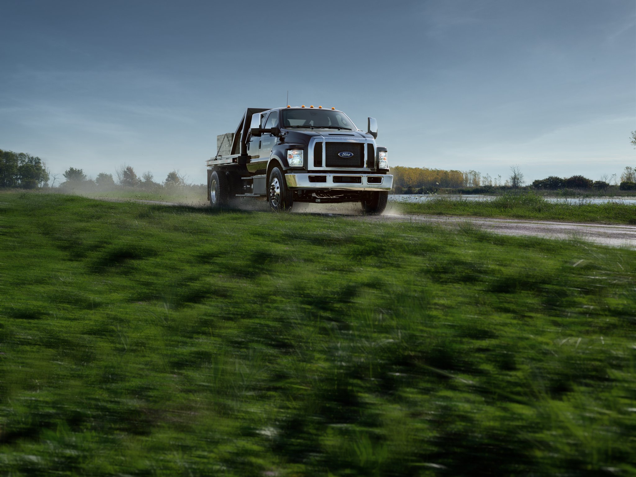 Ford F-750 2025 aménagé avec plateau à poteaux utilitaires en noir agate circulant sur un chemin de terre près de l’herbe et de l’eau