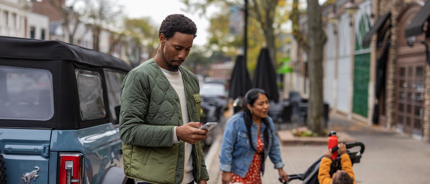A family stands on a sidewalk beside their parallel-parked Ford Bronco® while the father looks at his phone