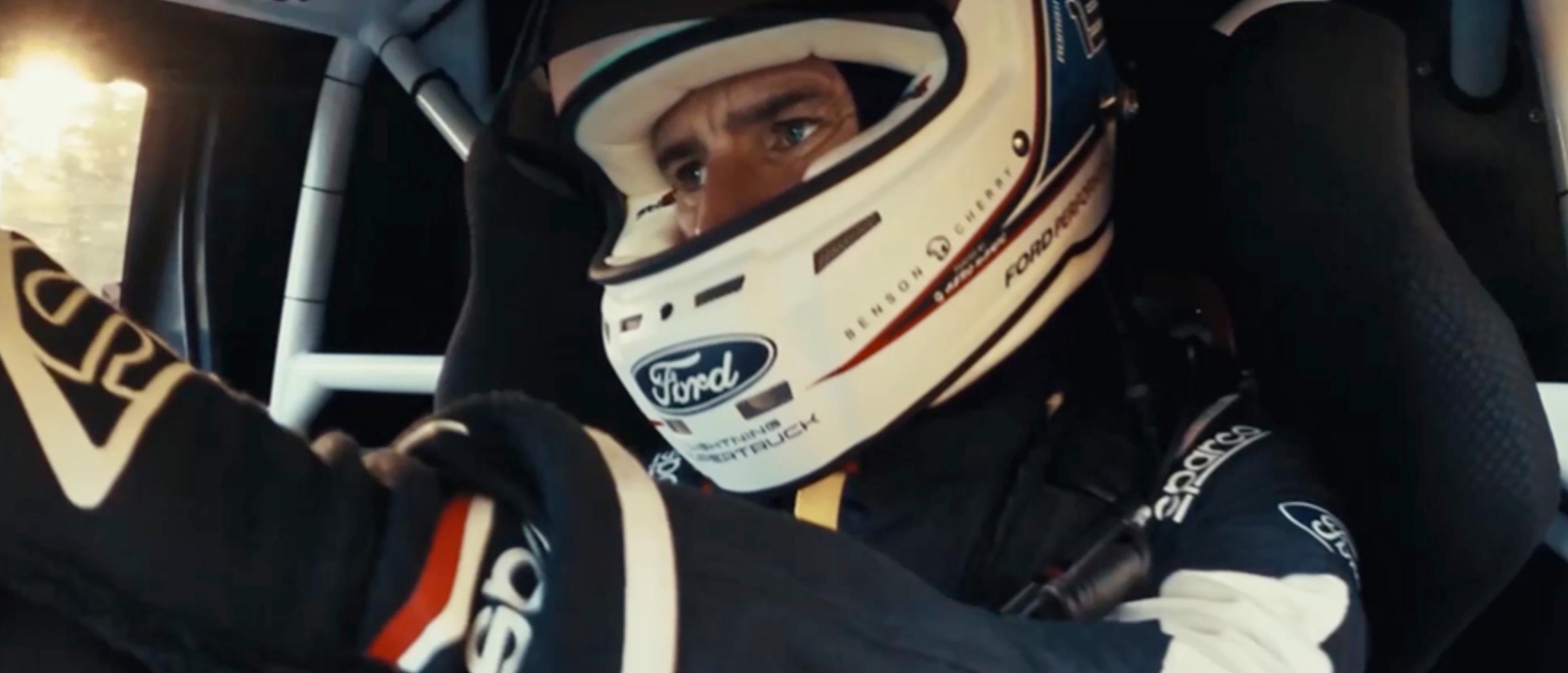 Ford race team member shown driving wearing flight suit and racing helmet