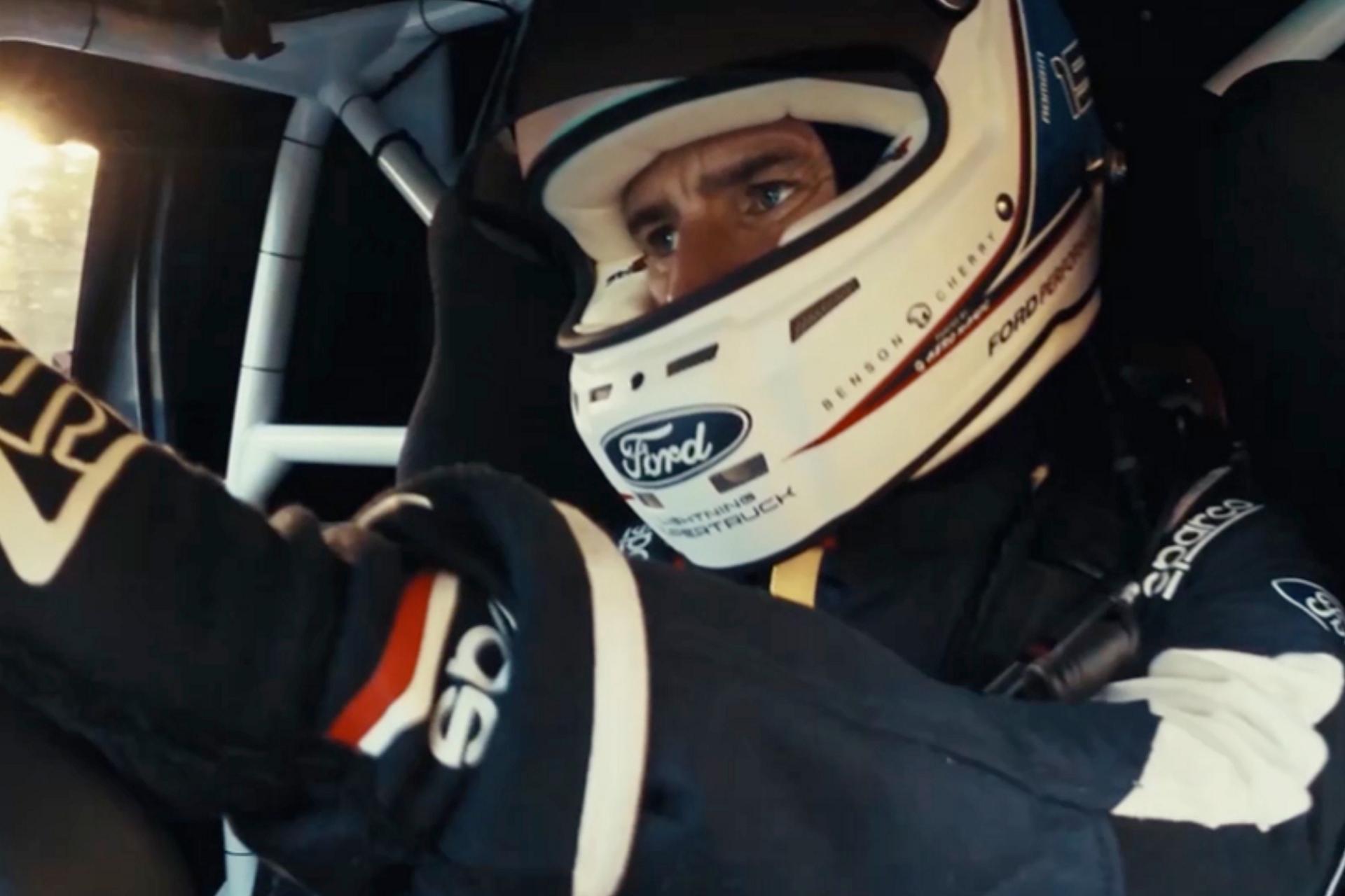 Ford race team member shown driving wearing flight suit and racing helmet