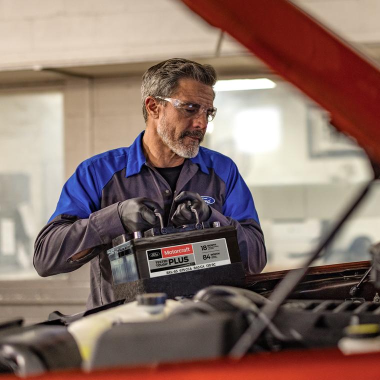 A Ford mechanic holding a battery near a Ford vehicle.  