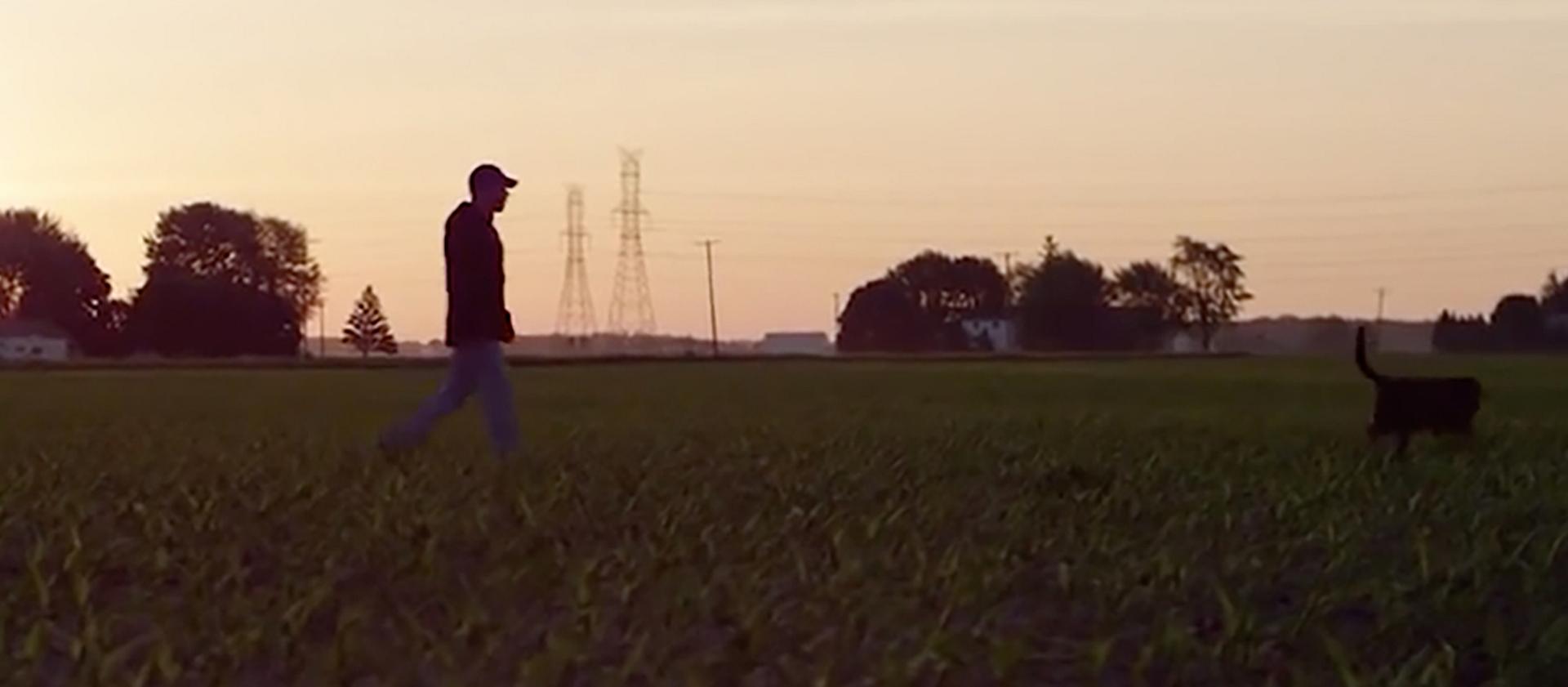 A farmer walks their dog across a wide open field at dusk