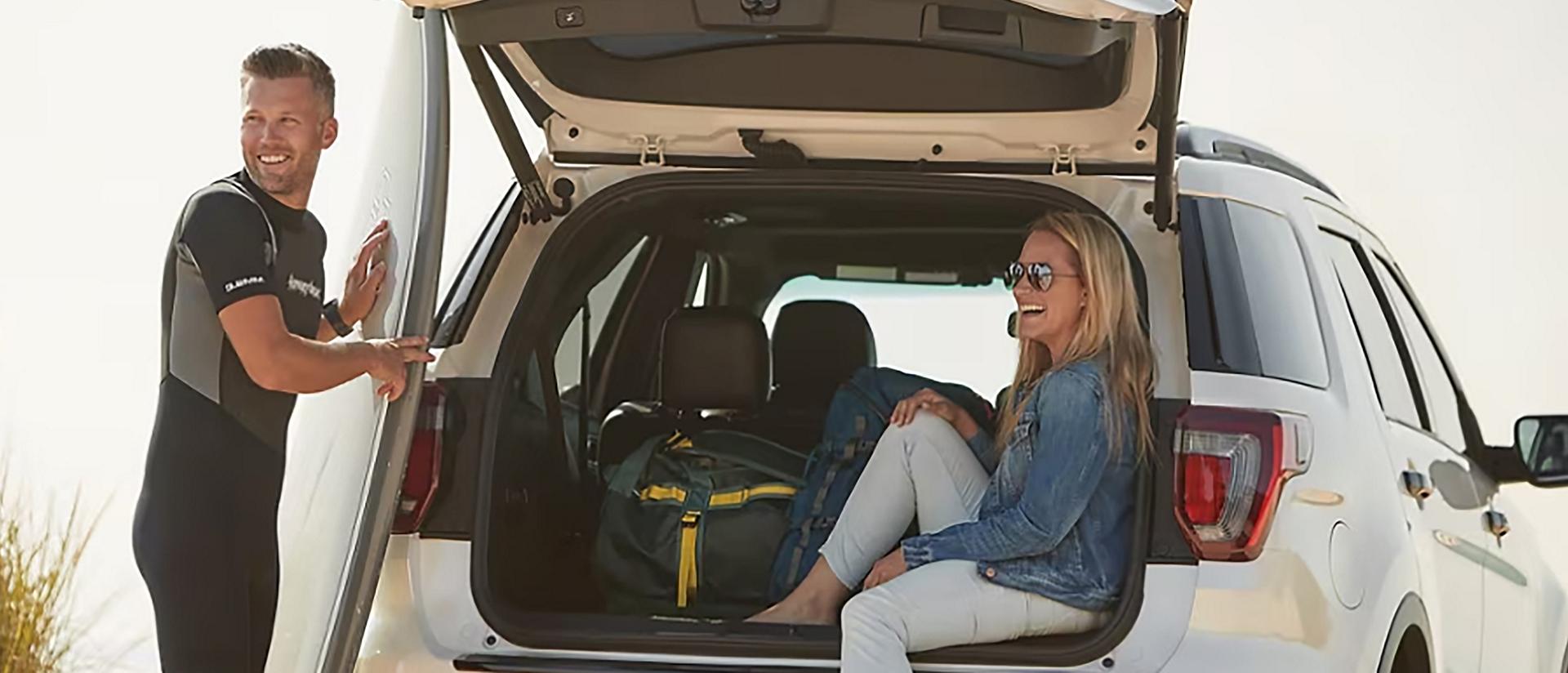 A person rests their surfboard against a Ford Explorer® SUV while another person sits inside under the open liftgate