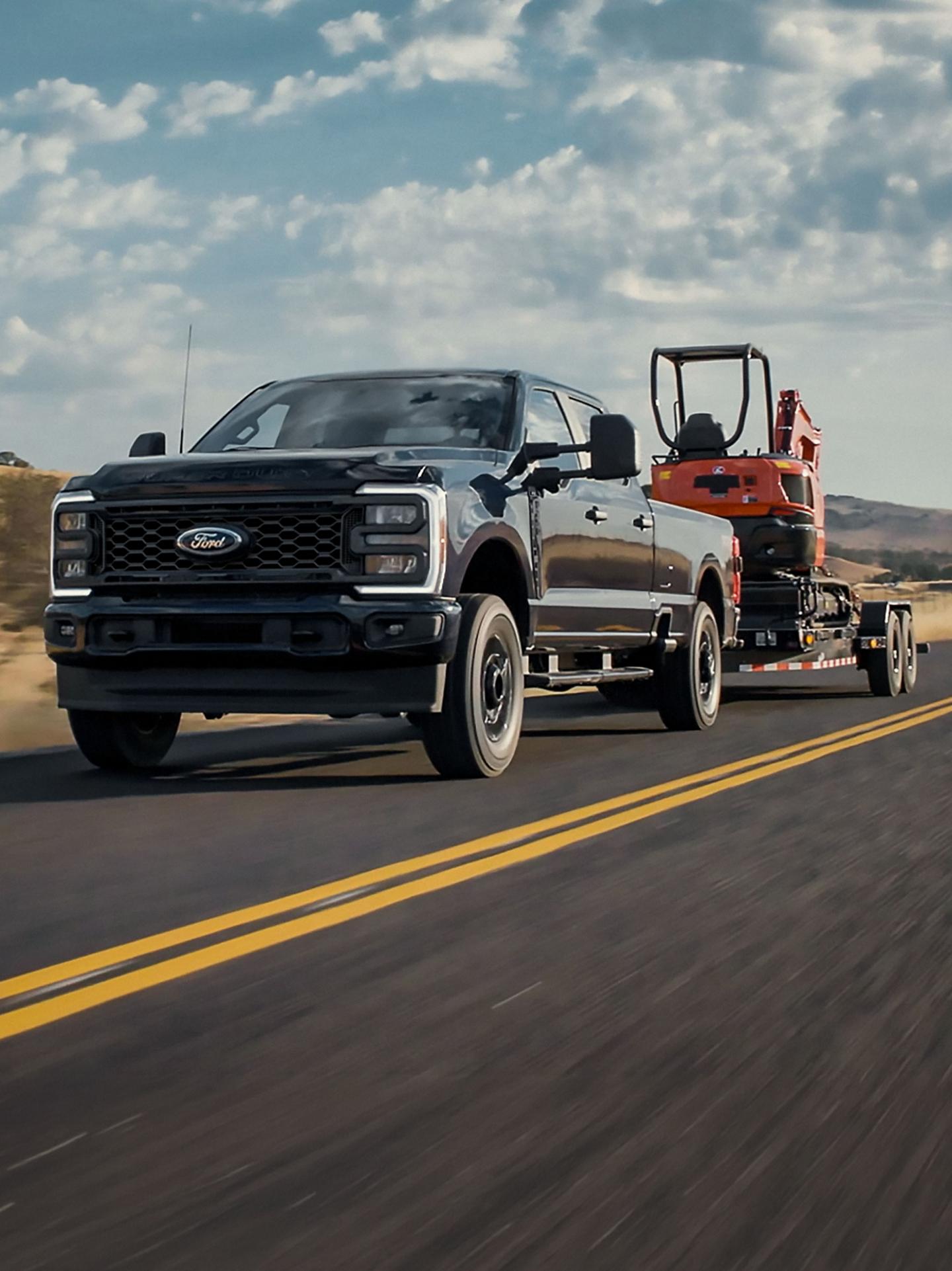 Imagen de una Camioneta black Super Duty Crew Cab remolcando un tráiler con una Miniexcavadora en el techo en una carretera de dos carriles.