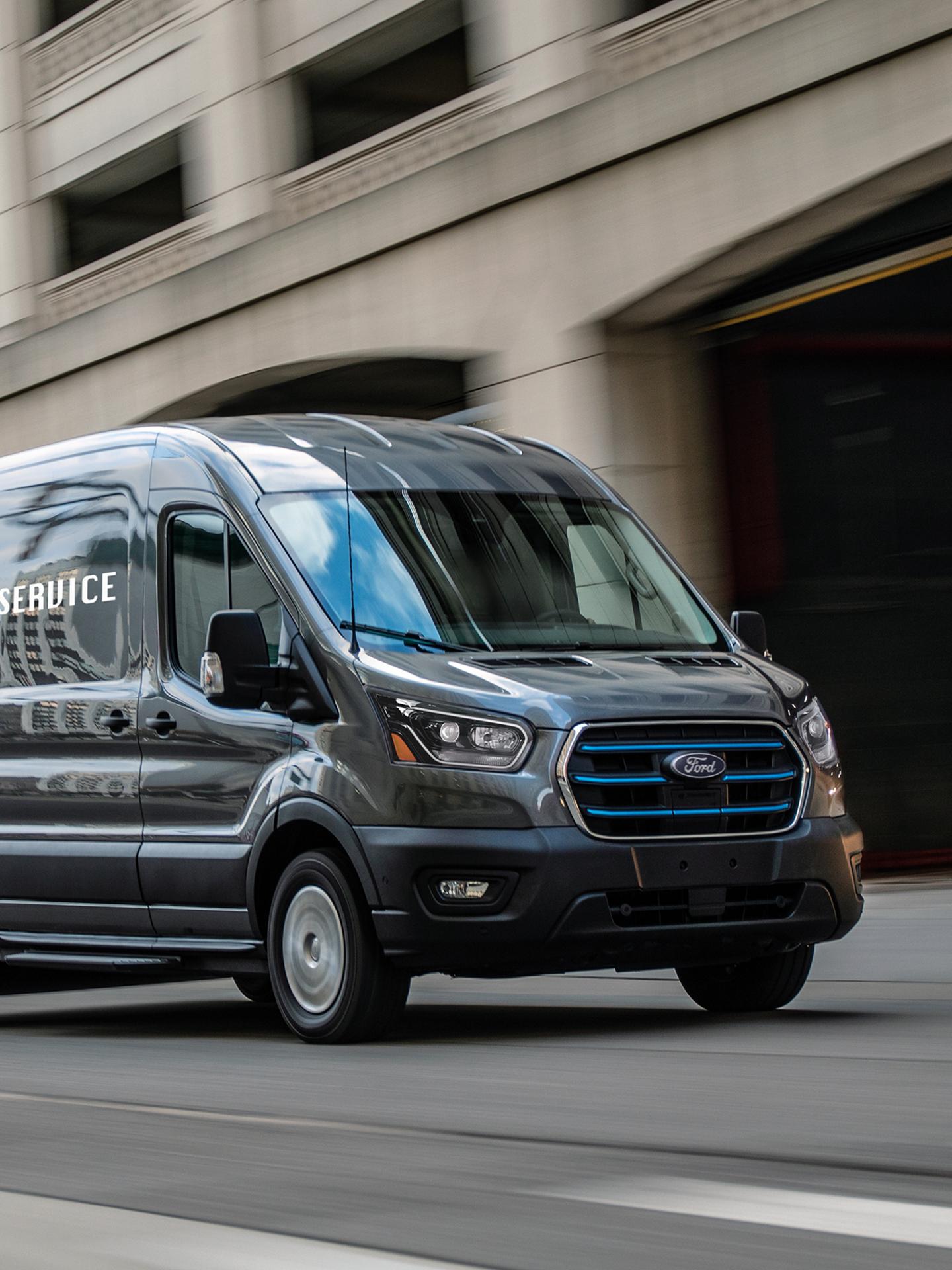 A dark grey 2027 Ford Transit® Van with blue grille accents driving on a city street alongside a modern building