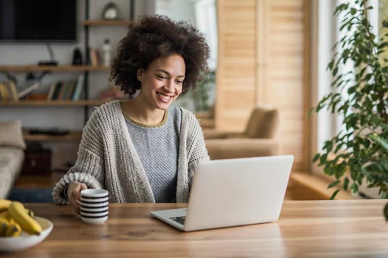 A person seated at a table in a cozy living space holds a coffee mug while looking at their laptop