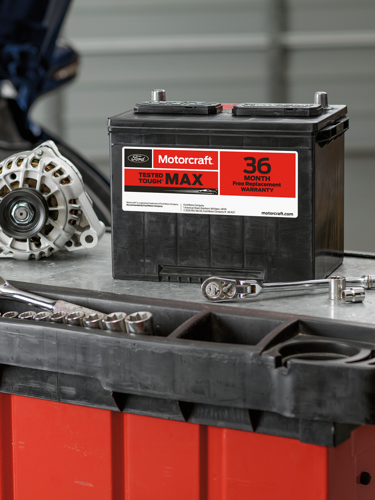 A Ford battery, wrench and vehicle parts sit on a workbench in front of a vehicle with its hood open inside a garage