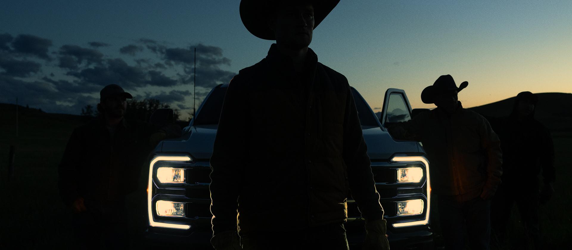 Silhouette of a person in a cowboy hat standing in front of a 2027 Super Duty® truck with the headlights on at dusk