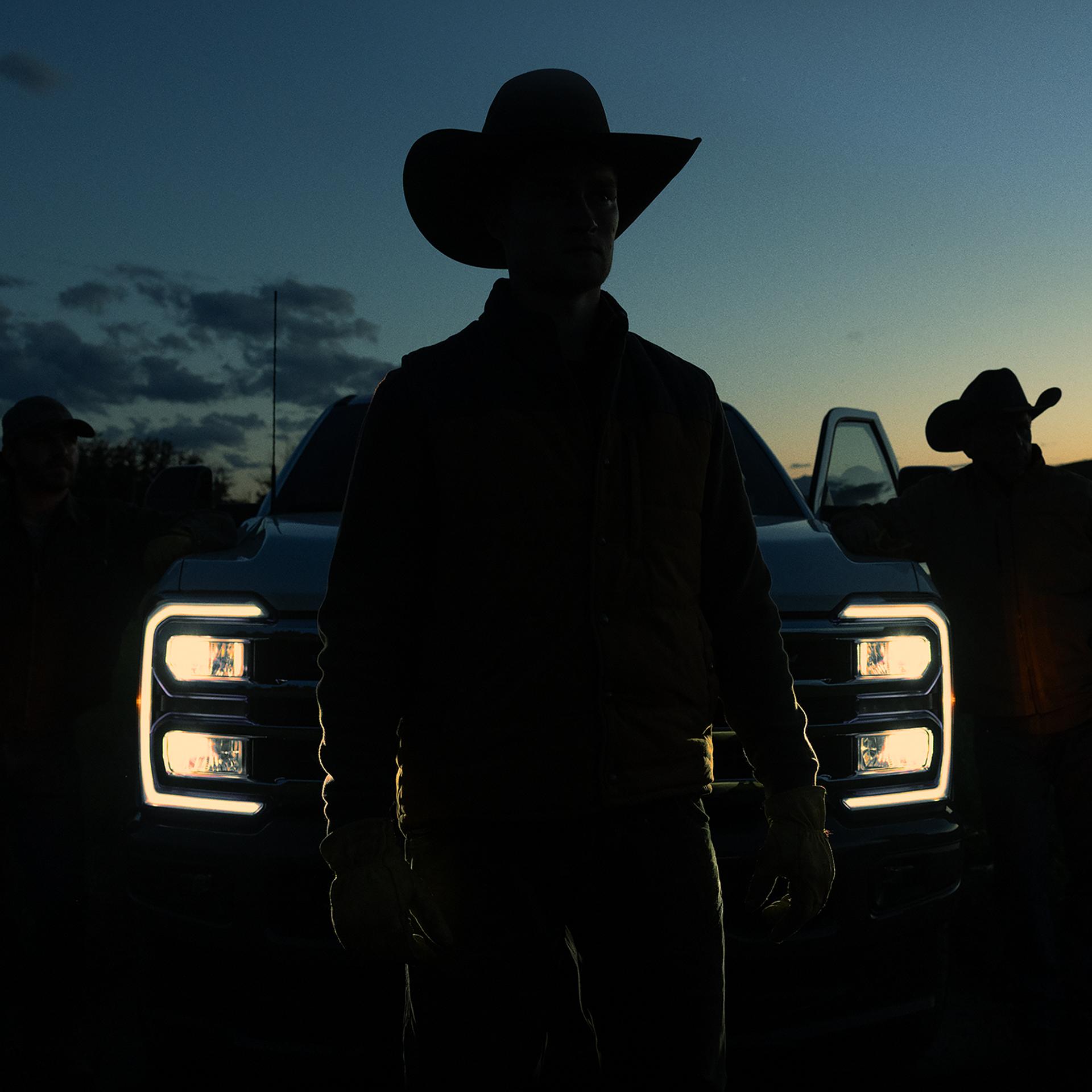 Silhouette of a person in a cowboy hat standing in front of a 2027 Super Duty® truck with the headlights on at dusk