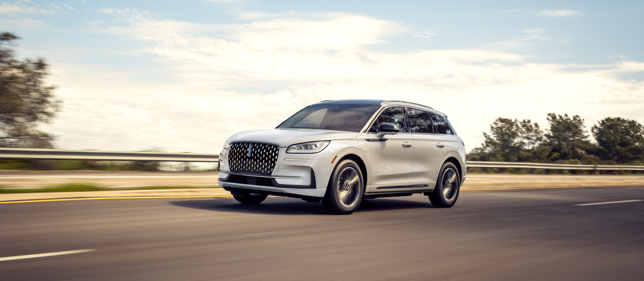 A 2024 Lincoln® Corsair Grand Touring in Pristine White is being driven on a freeway a blue sky and treetops in the background