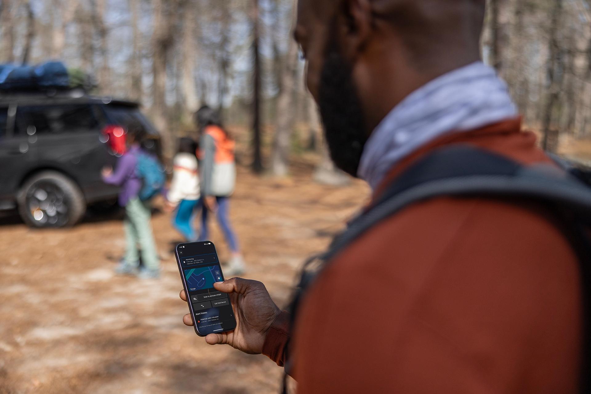 An adult looks at a cell phone while a family returns to a 2026 Ford Expedition® SUV parked in the woods in the distance