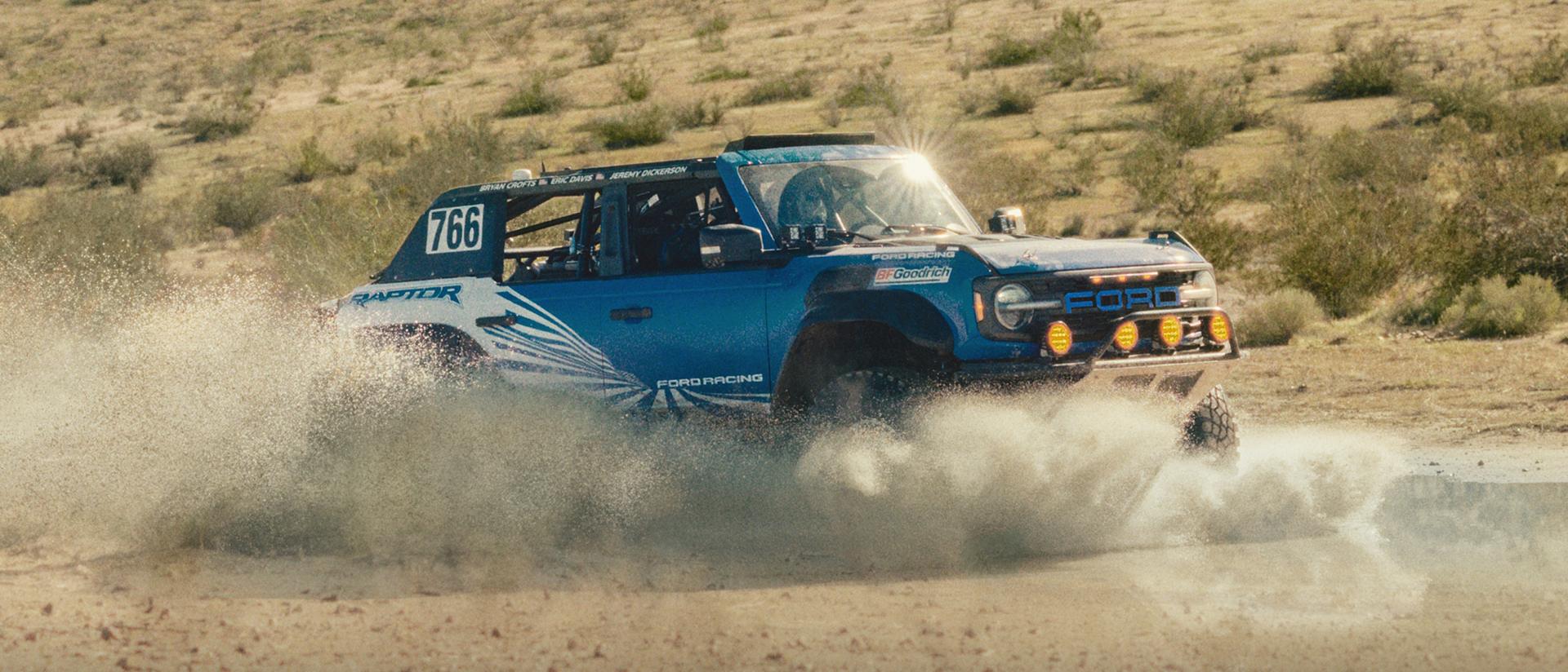 A blue with white graphics 2025 Ford Bronco® Raptor® racing through the desert