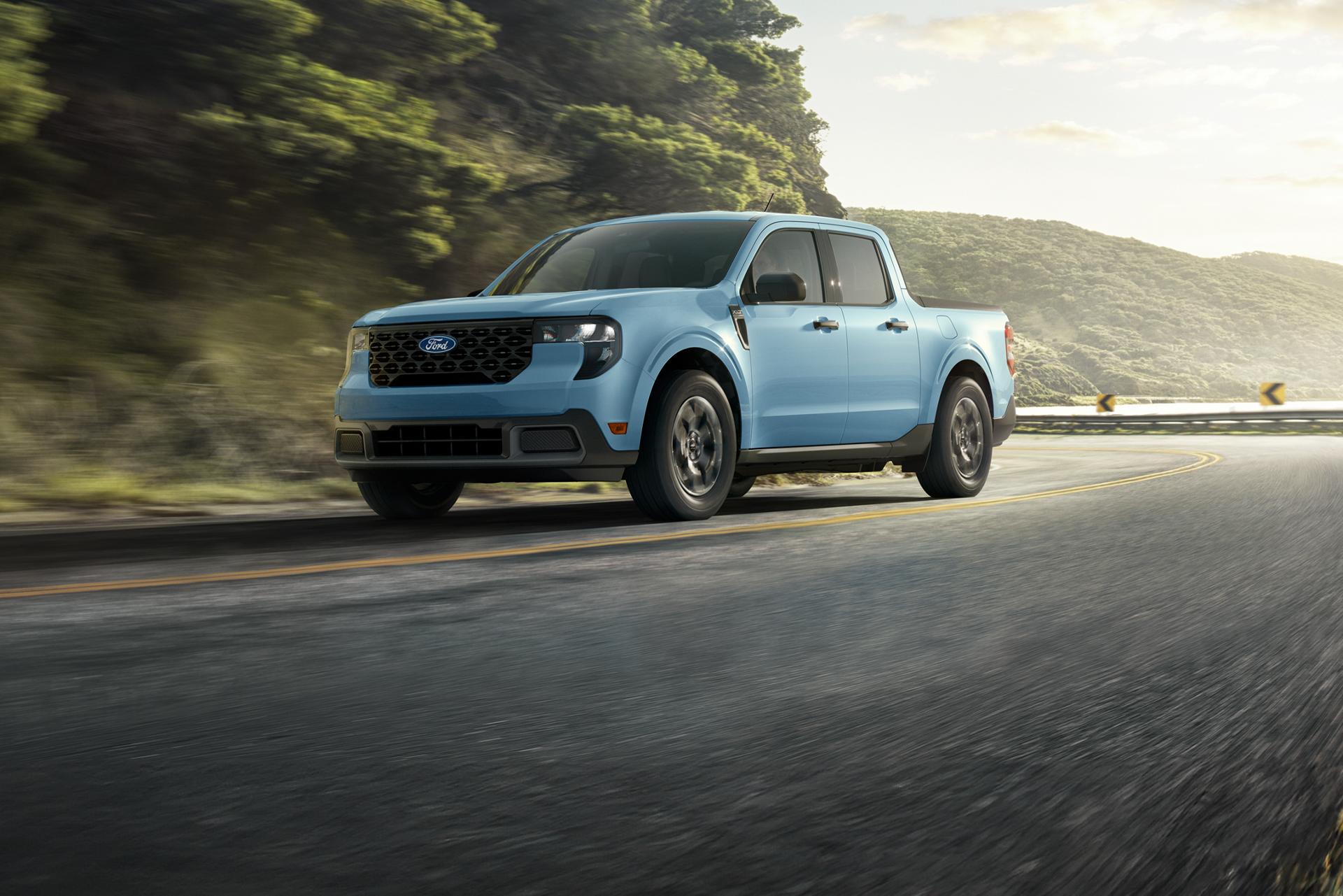 A gray 2026 Ford Maverick® pickup driving down a winding highway near a mountain lake