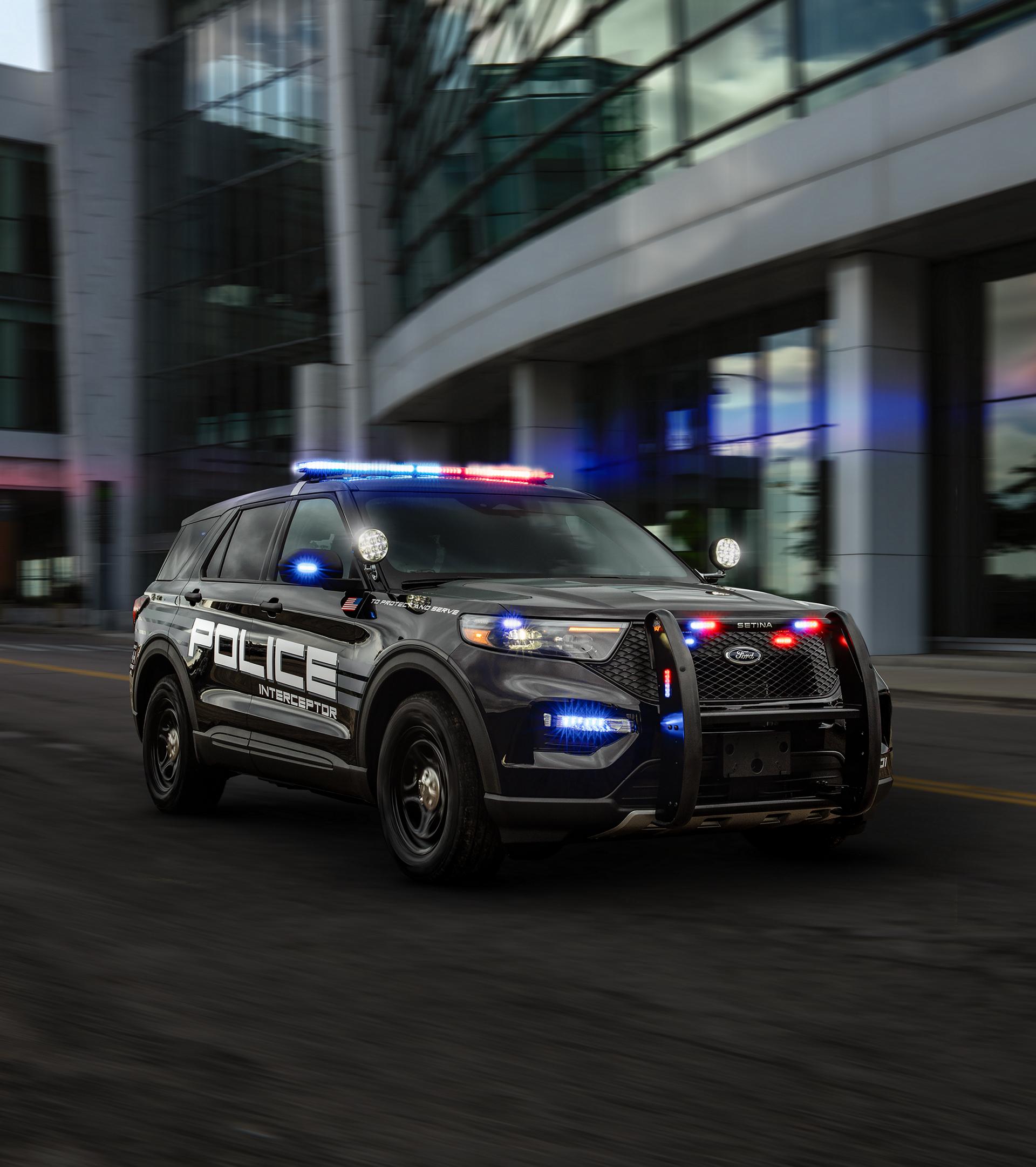 A Ford Explorer® Police Interceptor with lights on is driven on a city street past a building