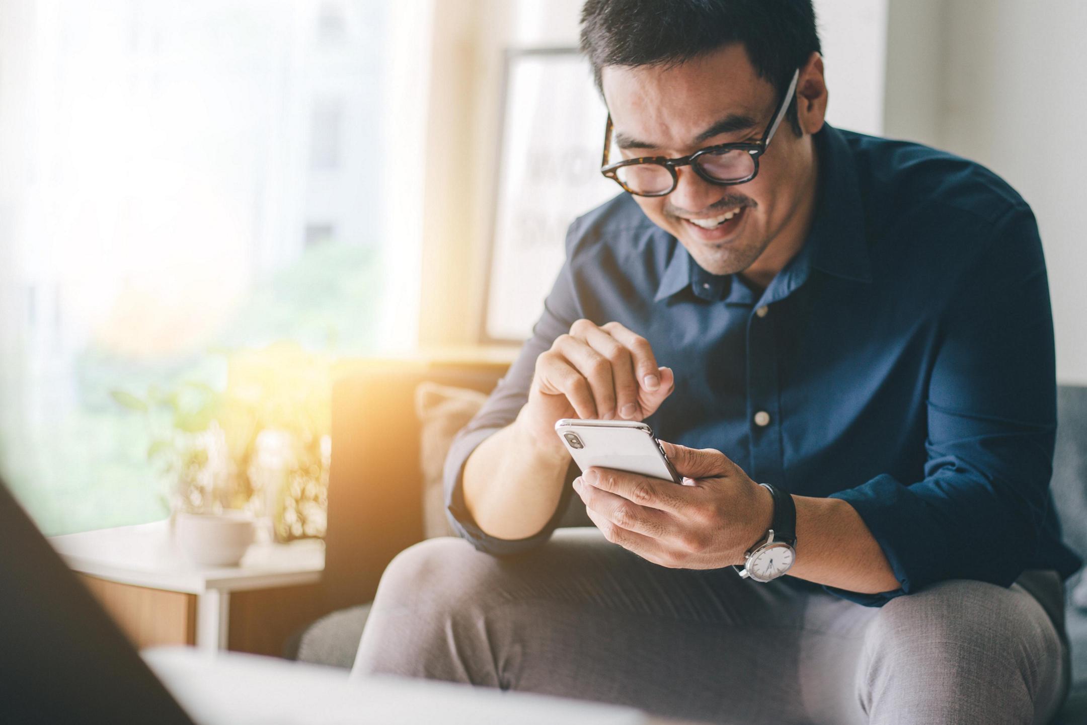 A man sitting on a couch in a sunny living room, smiling at his smartphone