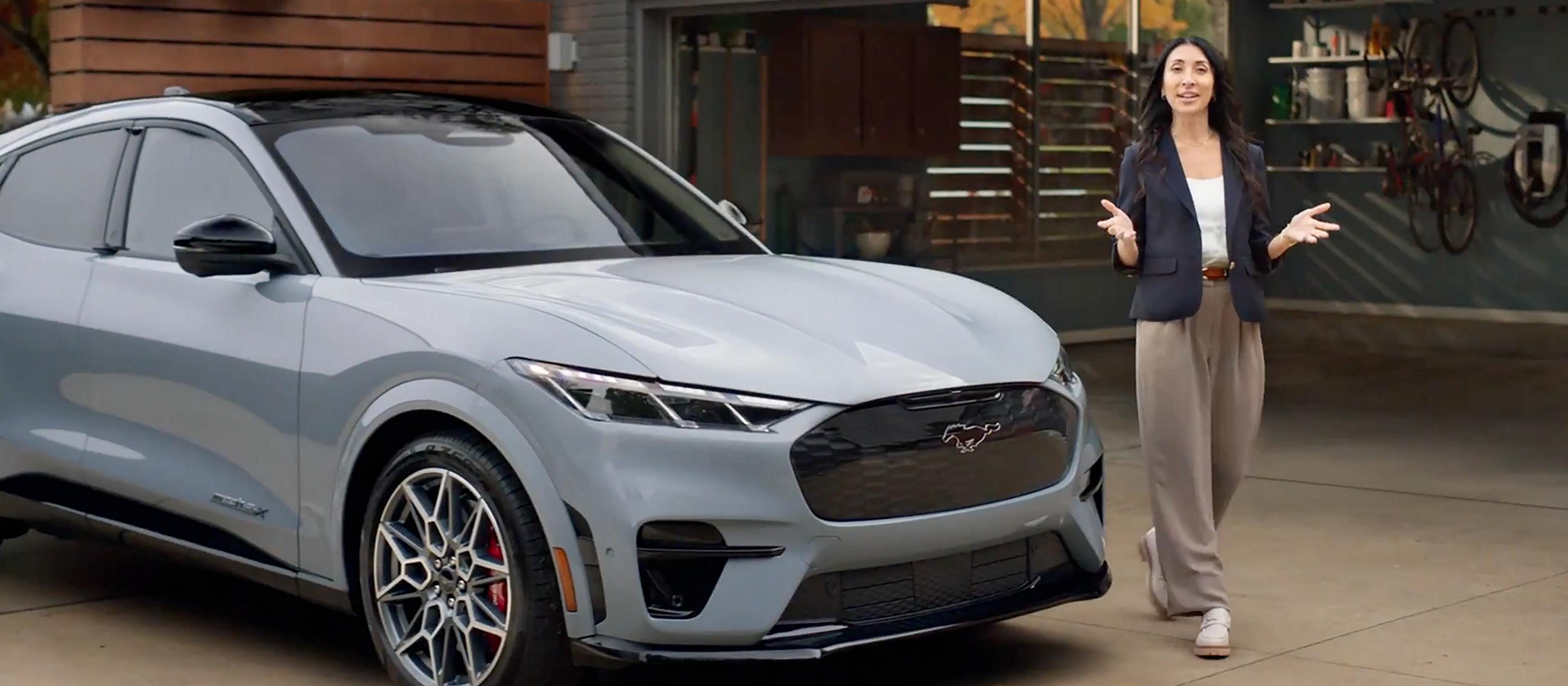 An adult walks around the front of a Ford Mustang Mach-E® SUV that is parked in front of a modern home