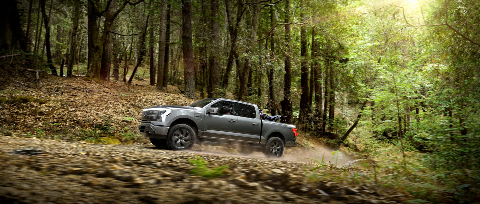 A 2025 Ford F-150® Lightning pickup driving on a dusty road through the forest with an ATV secured in the cargo bed