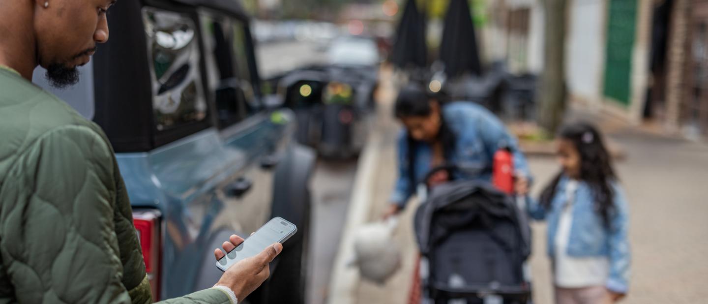 A family packs up a baby stroller next to their parallel-parked Ford Bronco® as the father looks at his phone
