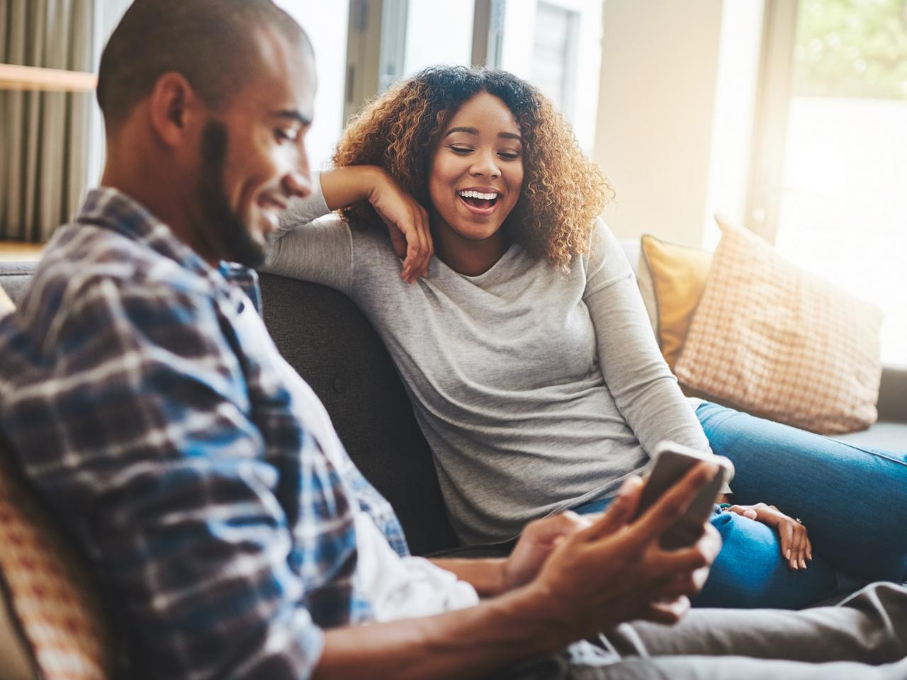 A couple sitting on a couch at home, smiling and looking at a smartphone together