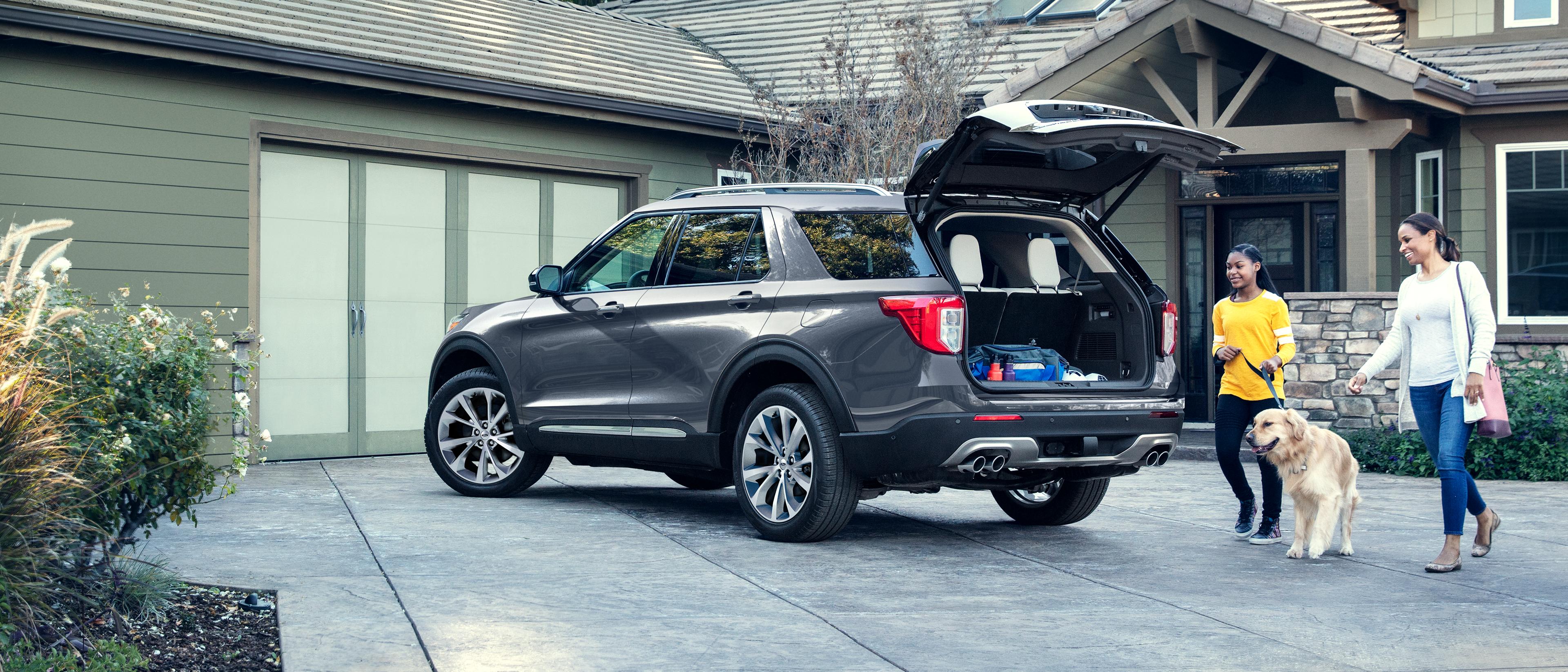 A couple of smiling people walk their dog over to the open liftgate of a Ford Explorer® SUV parked in the driveway