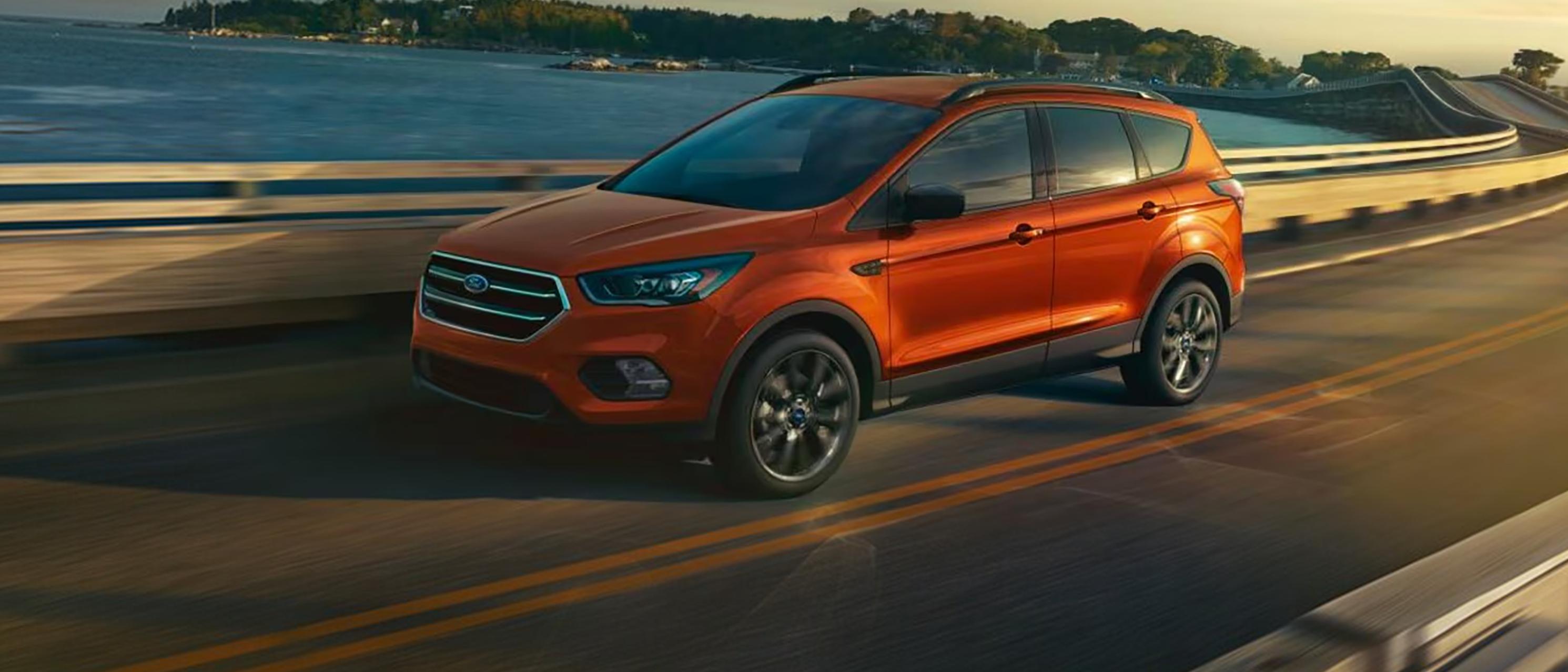 A Ford Escape® SUV is being driven over a bridge along a body of water at sunset