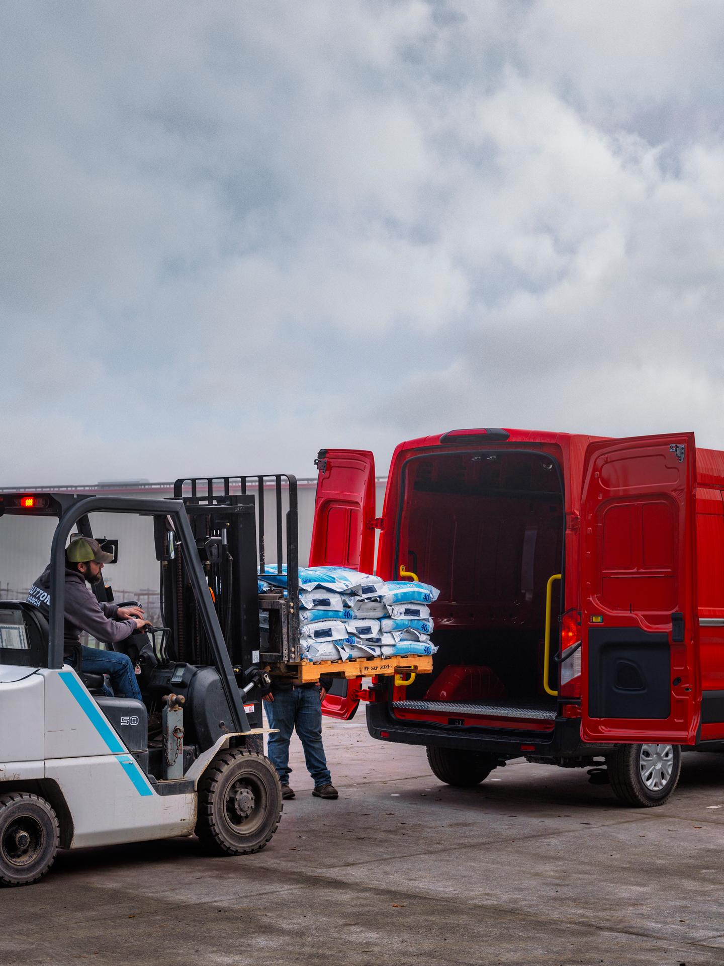 A forklift loads sacks into the back of an open red 2027 Ford Transit® Cargo van, parked outside a building