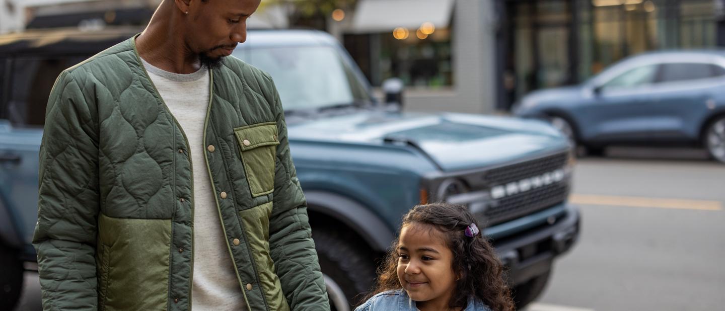 A father and daughter walk hand-in-hand on a sidewalk with their parked Ford Bronco® in the background