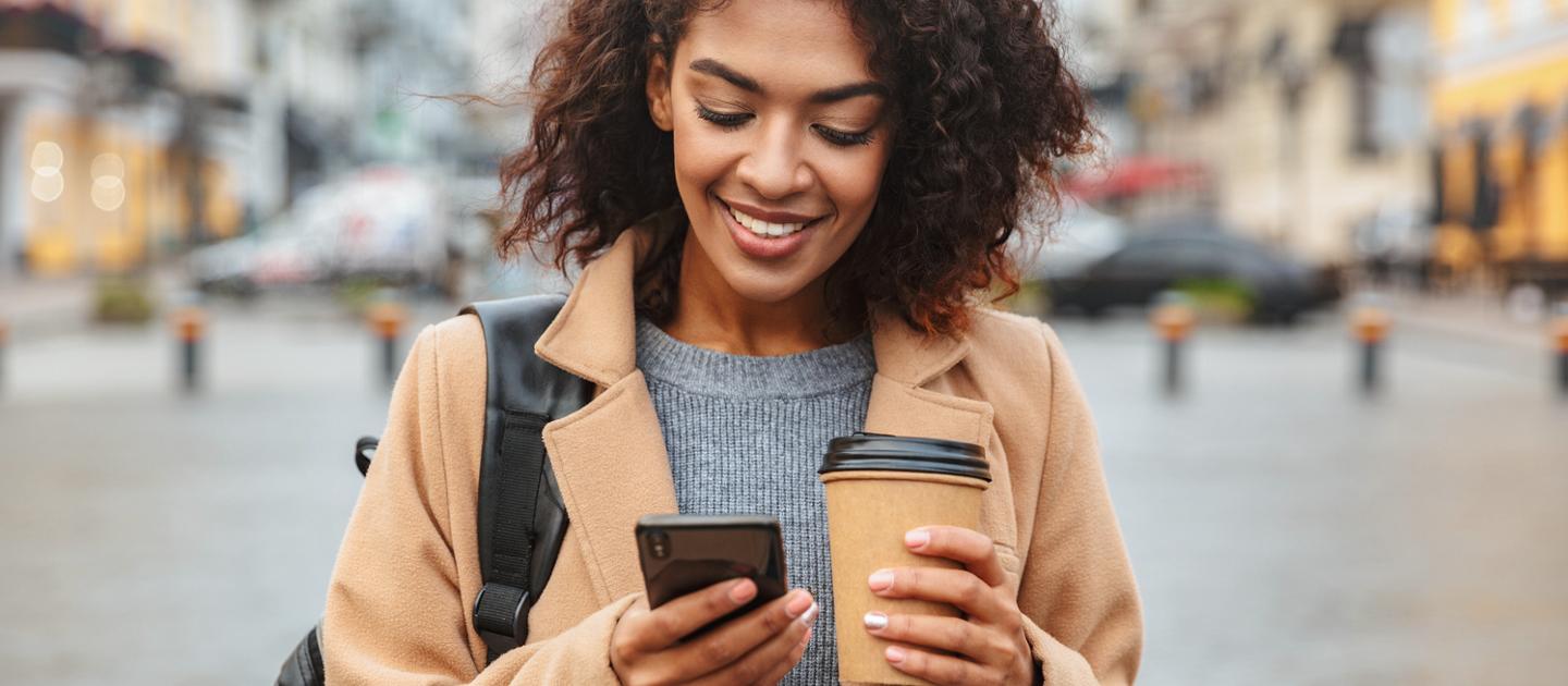 Person standing outside holding their smartphone in one hand and a coffee in the other