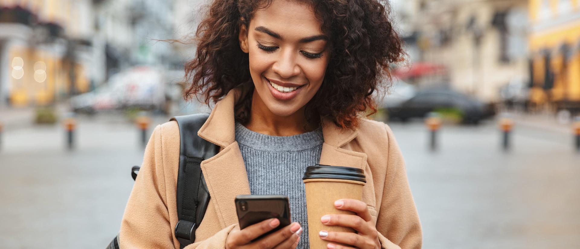 Person standing outside holding their smartphone in one hand and a coffee in the other