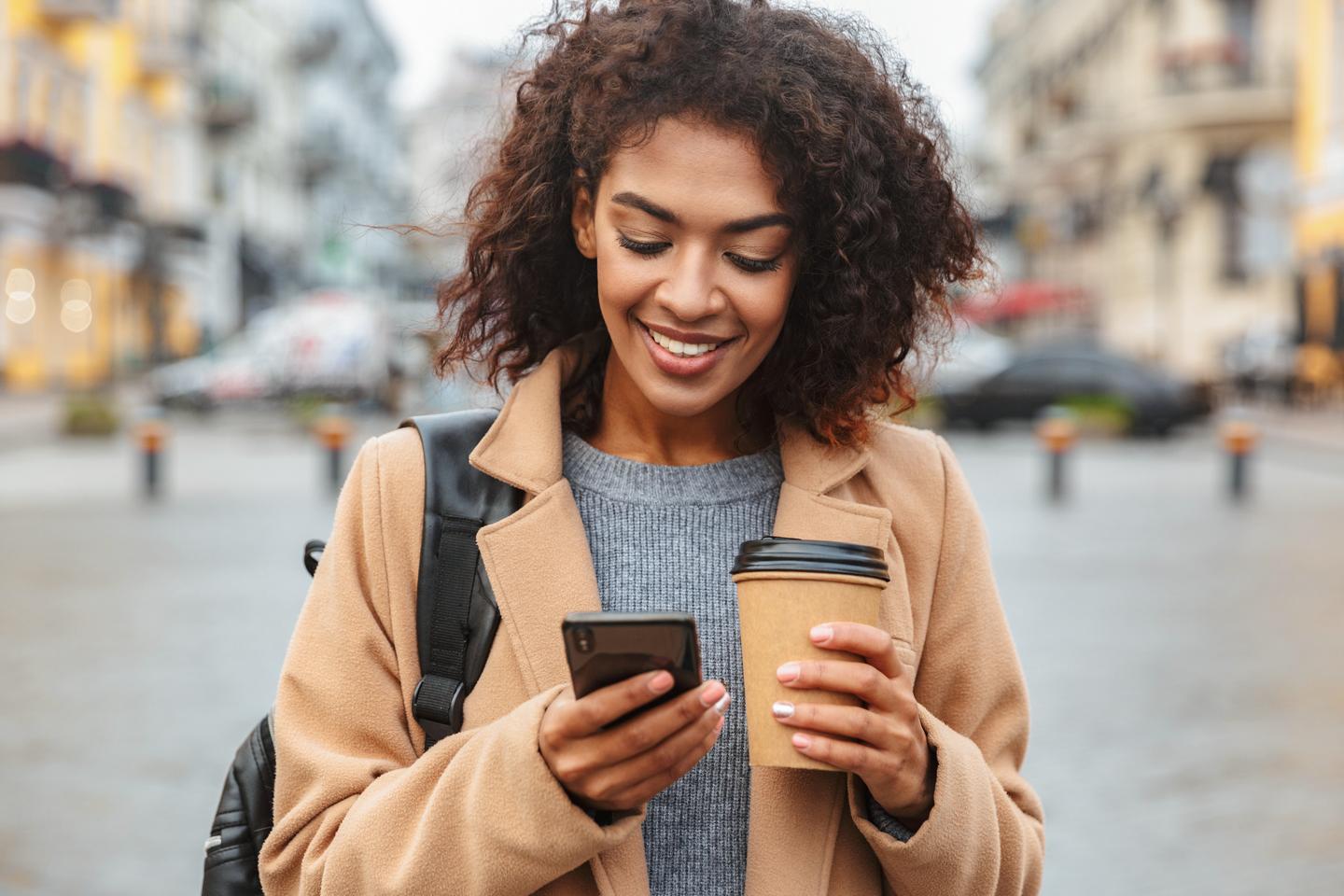 Person standing outside holding their smartphone in one hand and a coffee in the other