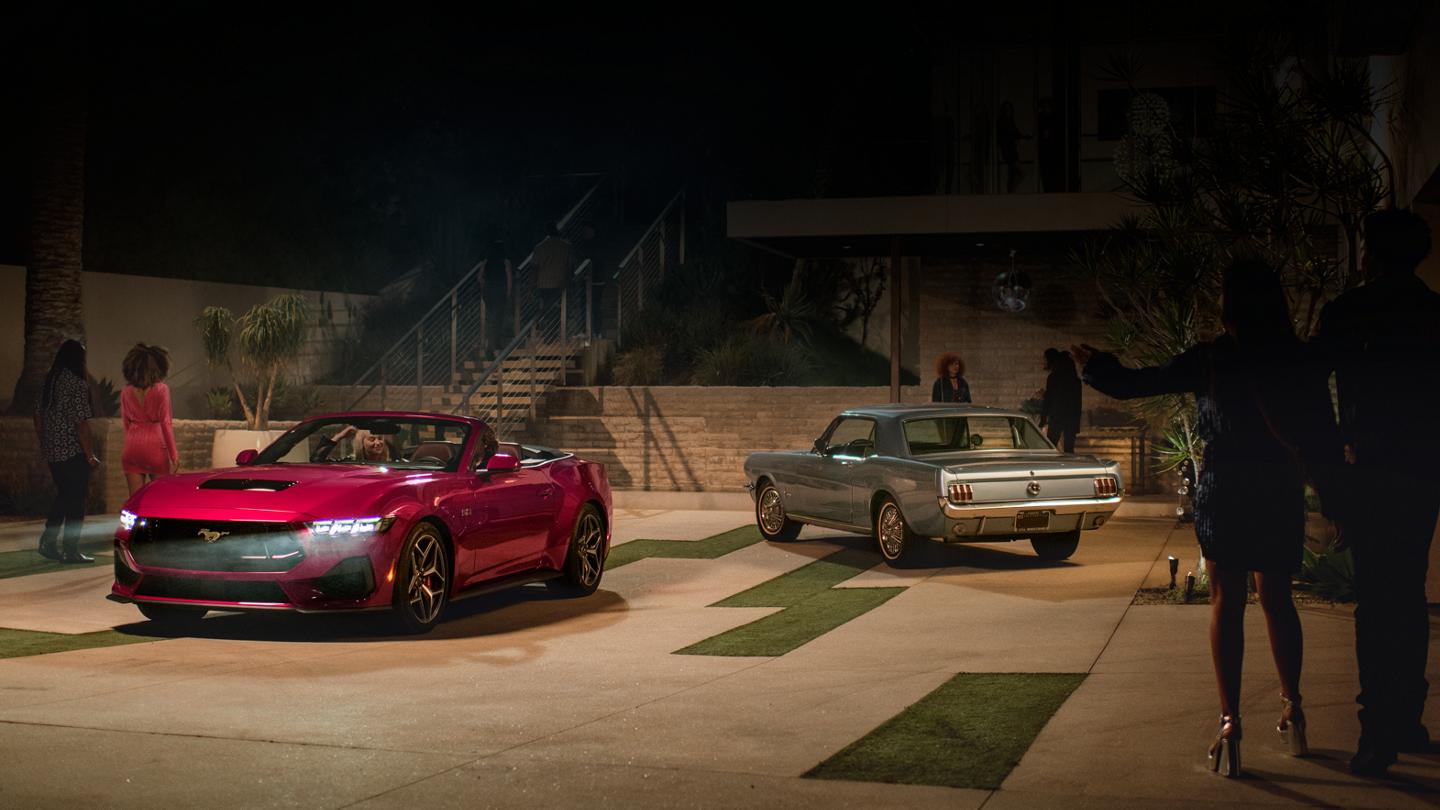2025 Mustang® GT convertible and a first-gen Mustang coupe parked in a driveway with people nearby