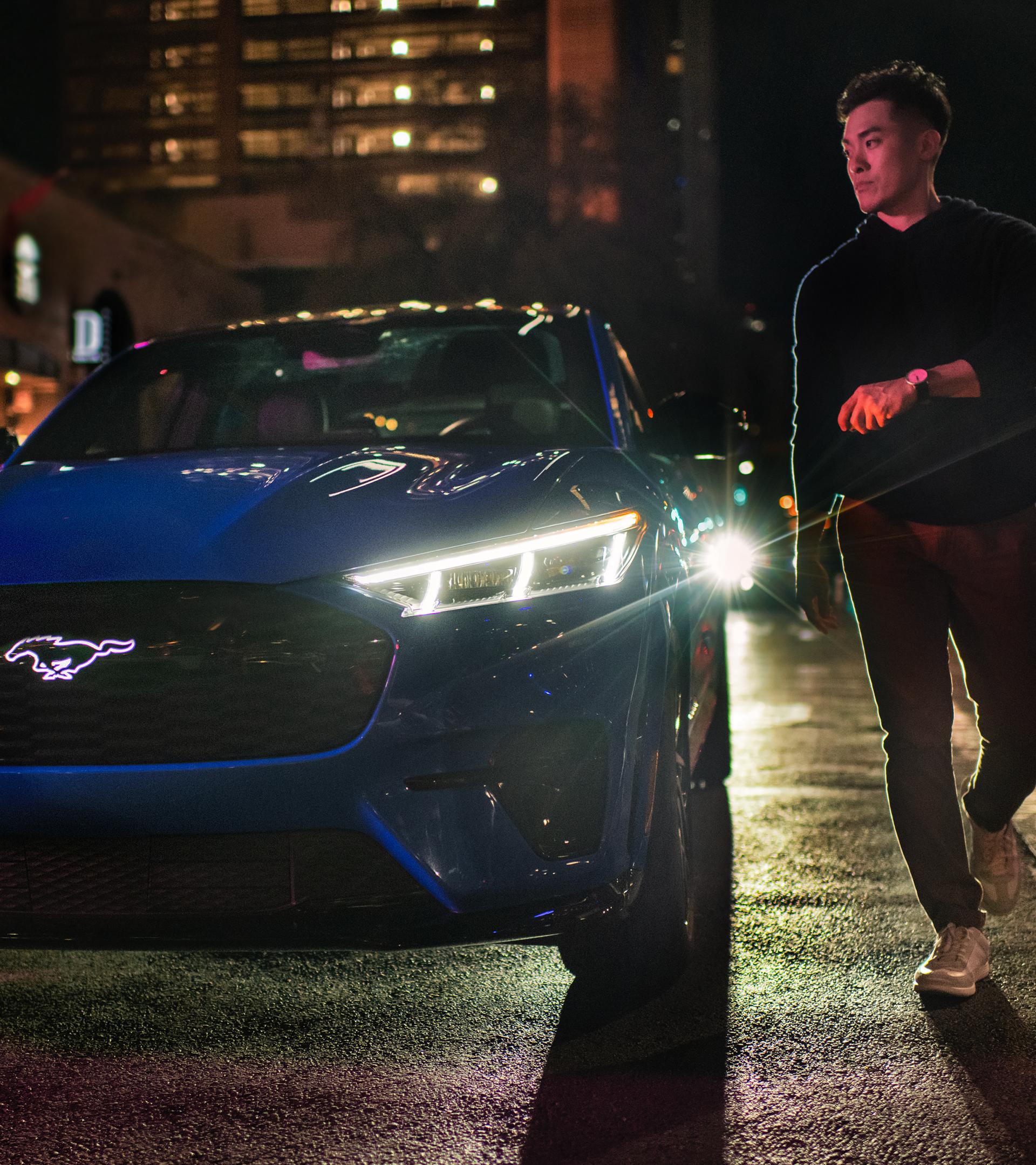 A Ford Mustang Mach-E® SUV owner walks around the front of the vehicle after parking on a city street