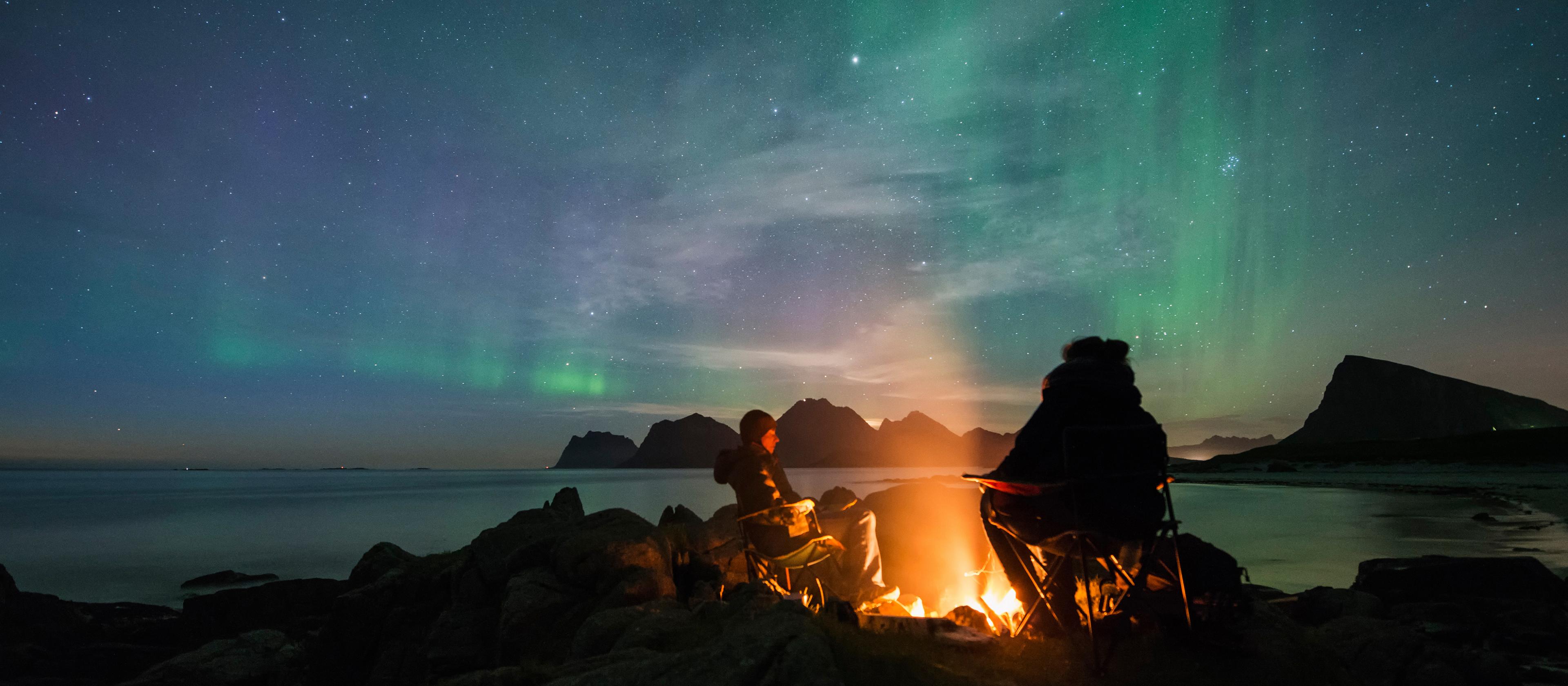 A couple enjoy a campfire while the Northern Lights dance in the sky above