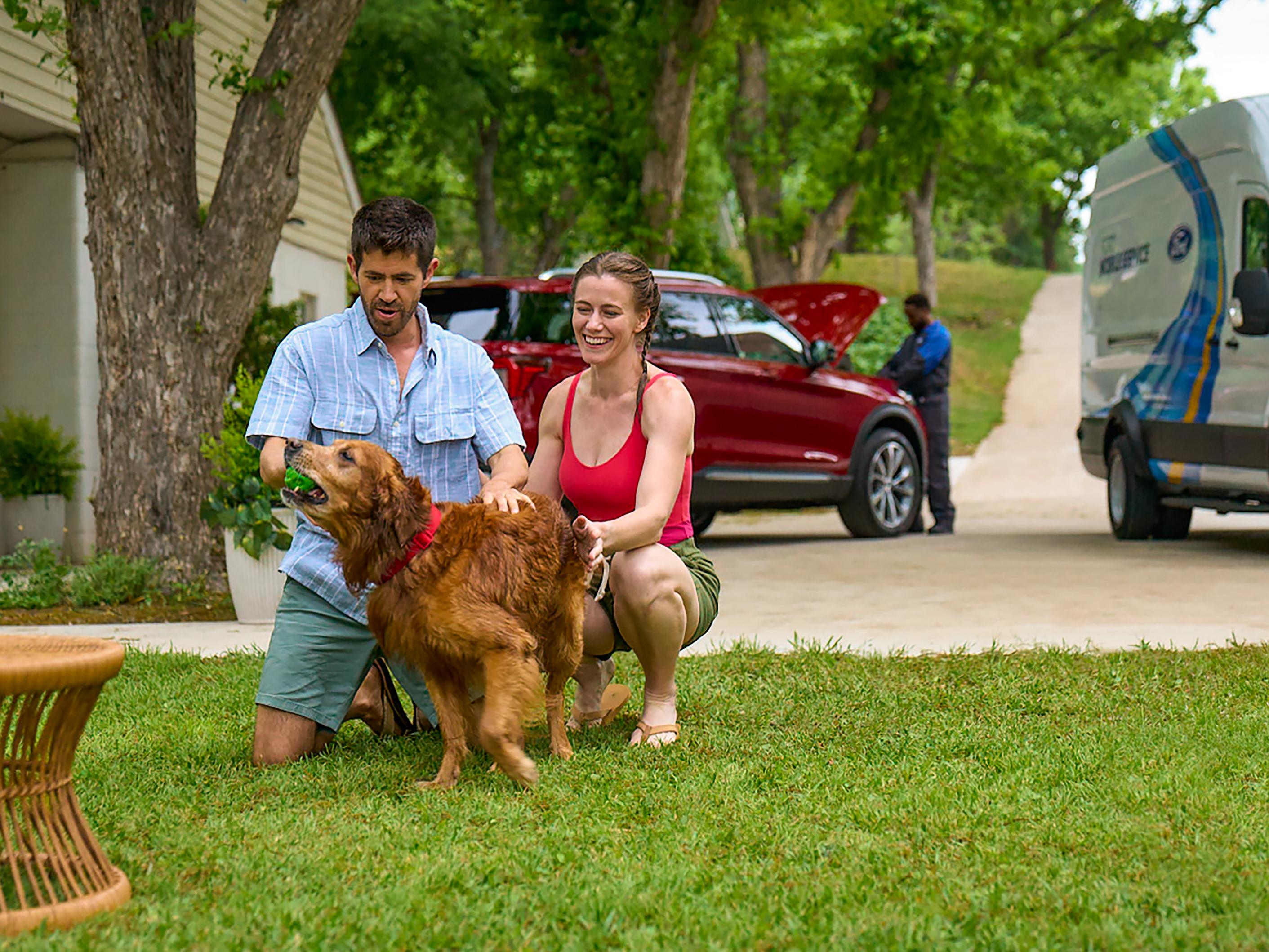 Two adults play with their dog on their front lawn while a Ford technician works on their Ford vehicle in the background