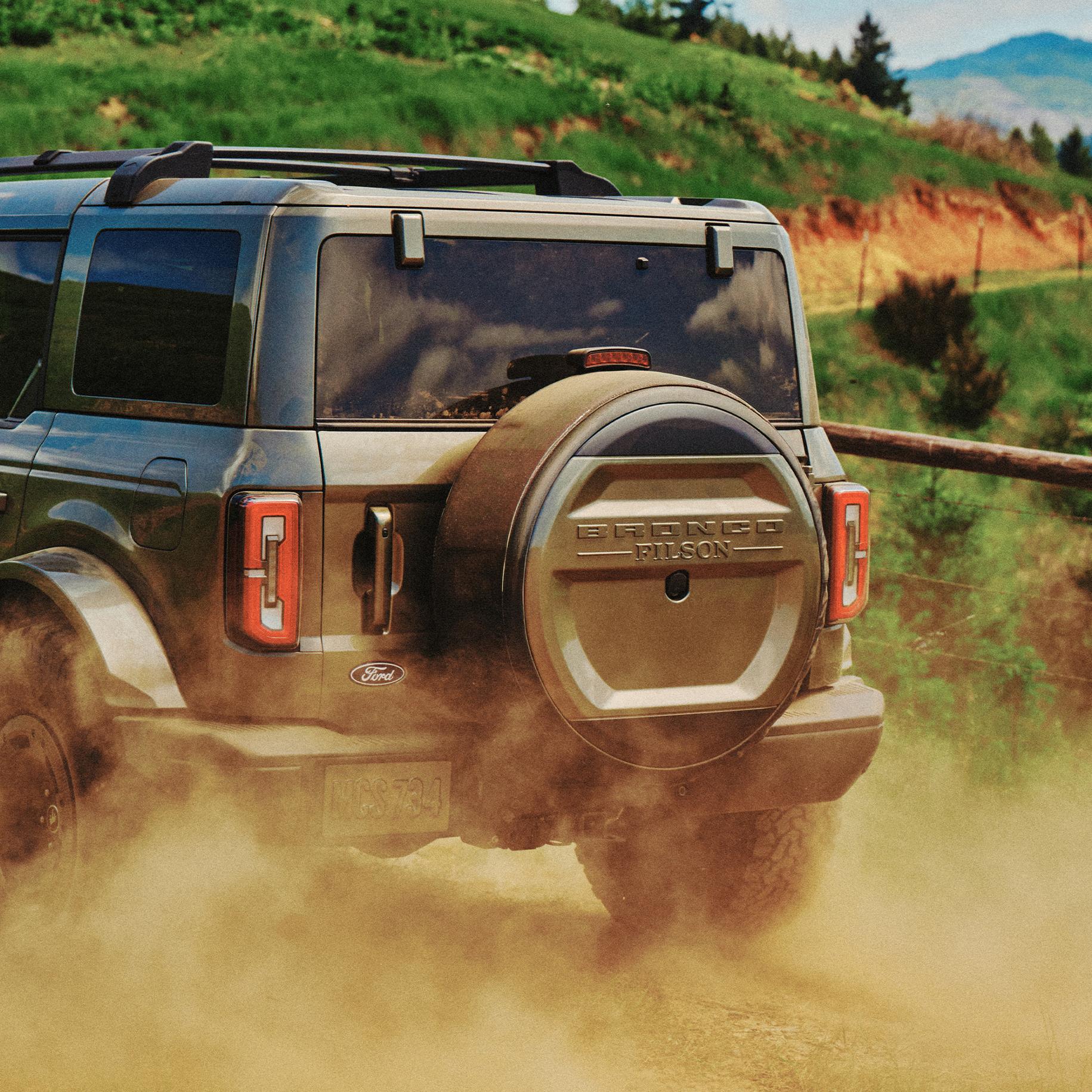 A view from behind a 2027 Ford Bronco® Filson™ SUV as it kicks up dust on a dirt trail