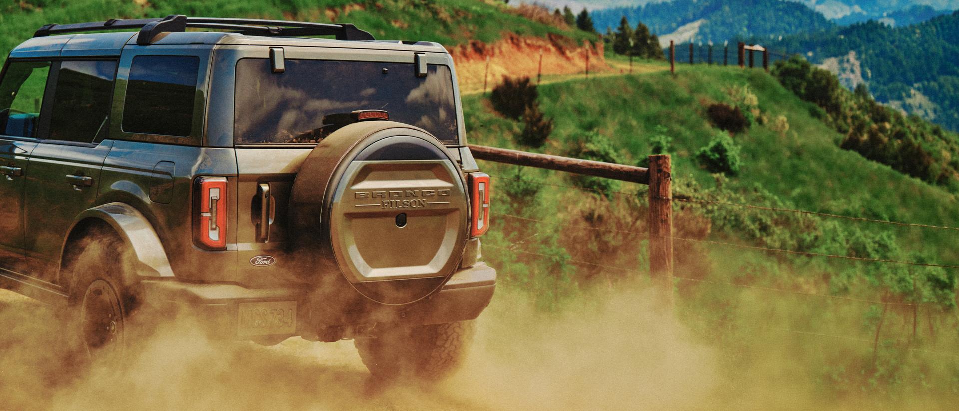 A view from behind a 2027 Ford Bronco® Filson™ SUV as it kicks up dust on a dirt trail
