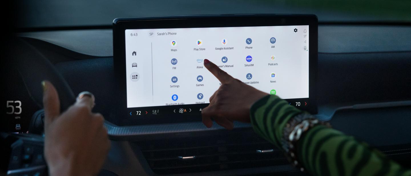 Someone drives a Ford vehicle while a passenger selects Alexa from a library of options on the vehicle’s center screen.