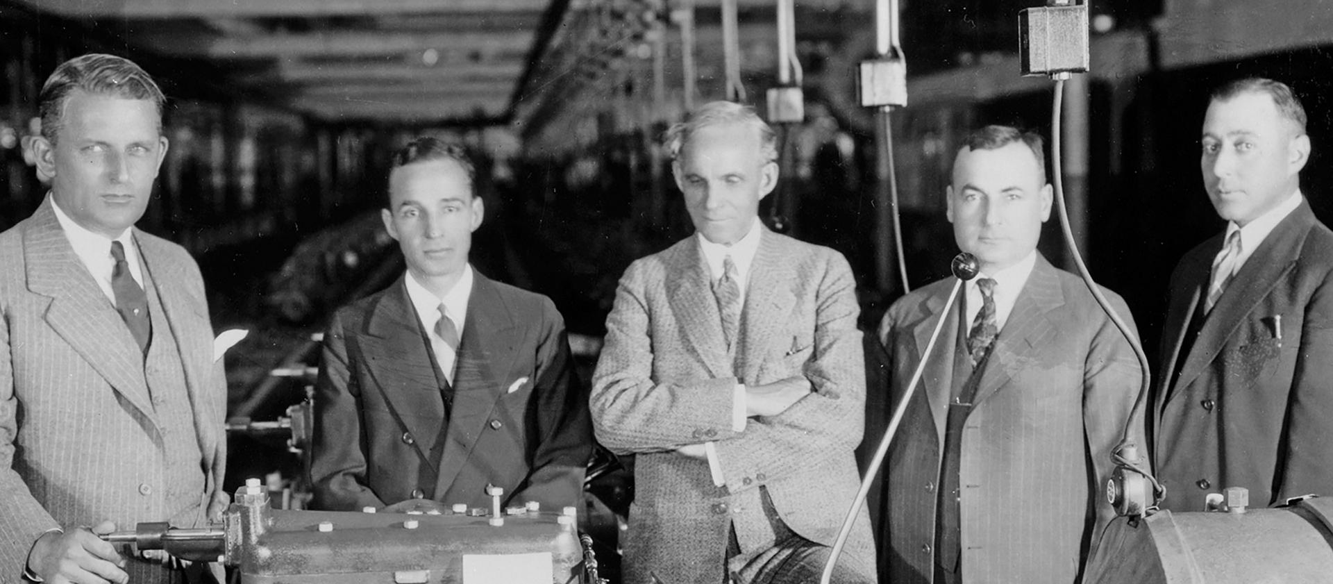 A black and white photo of Henry Ford lined up with executives inspecting a Model A engine